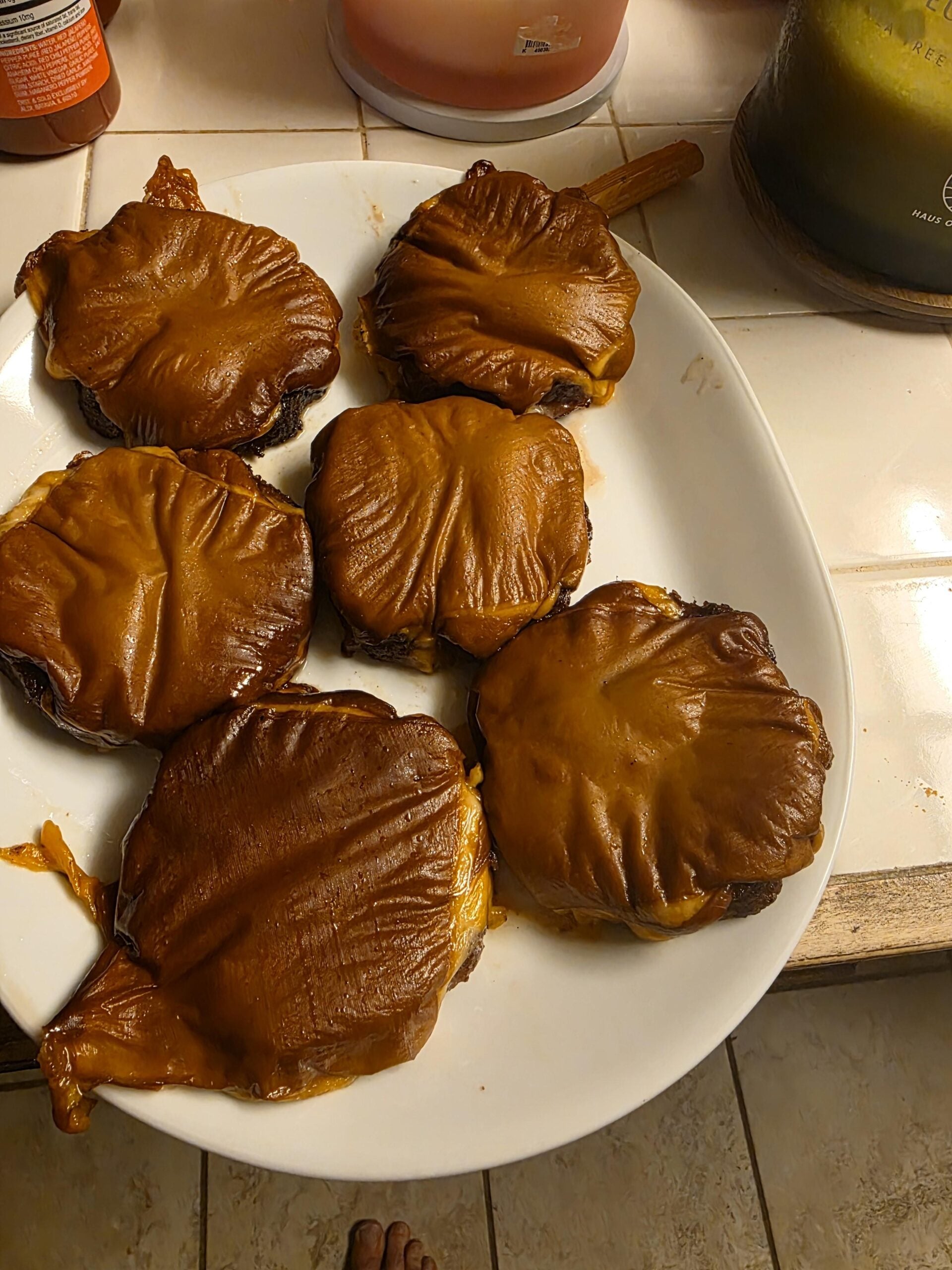 Six burgers topped with melted cheddar cheese on a white plate, sitting on a tiled kitchen counter.
