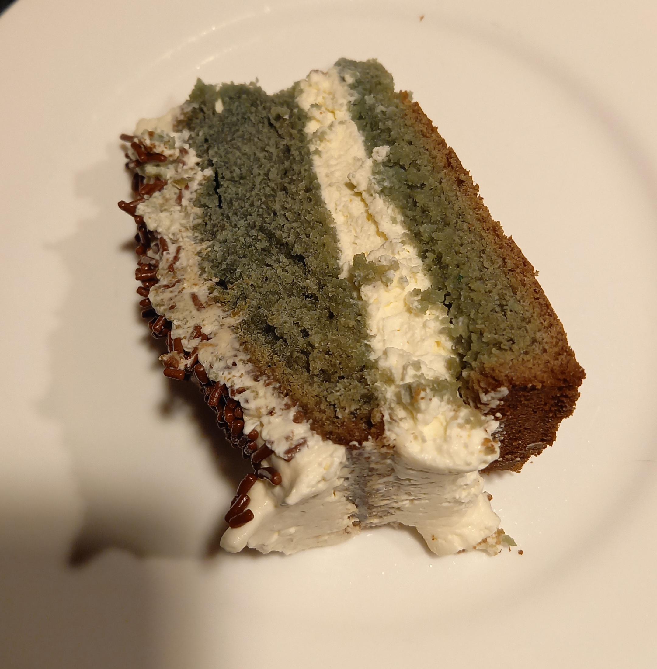 A slice of greenish-brown cake with white cream filling and frosting sits on a white plate.