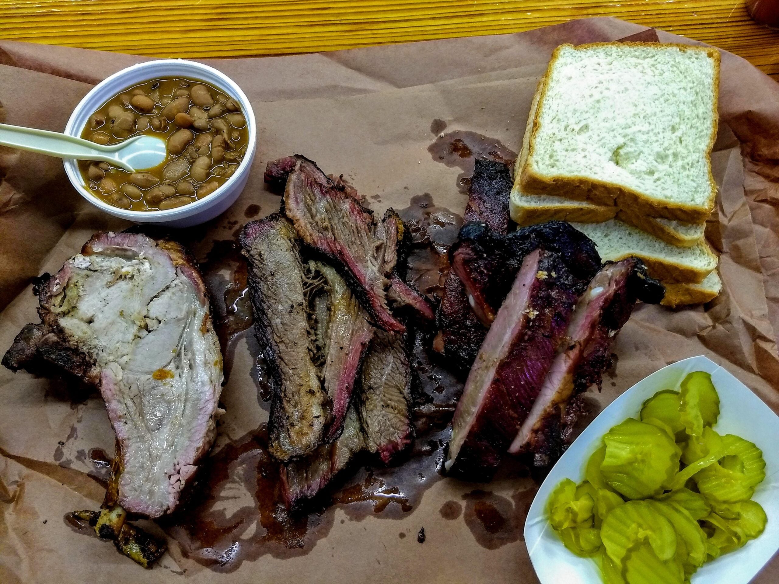 Barbecue plate with smoked brisket, pork ribs, sliced bread, a small bowl of baked beans with a spoon, and a side of sliced pickles on a sheet of brown paper.