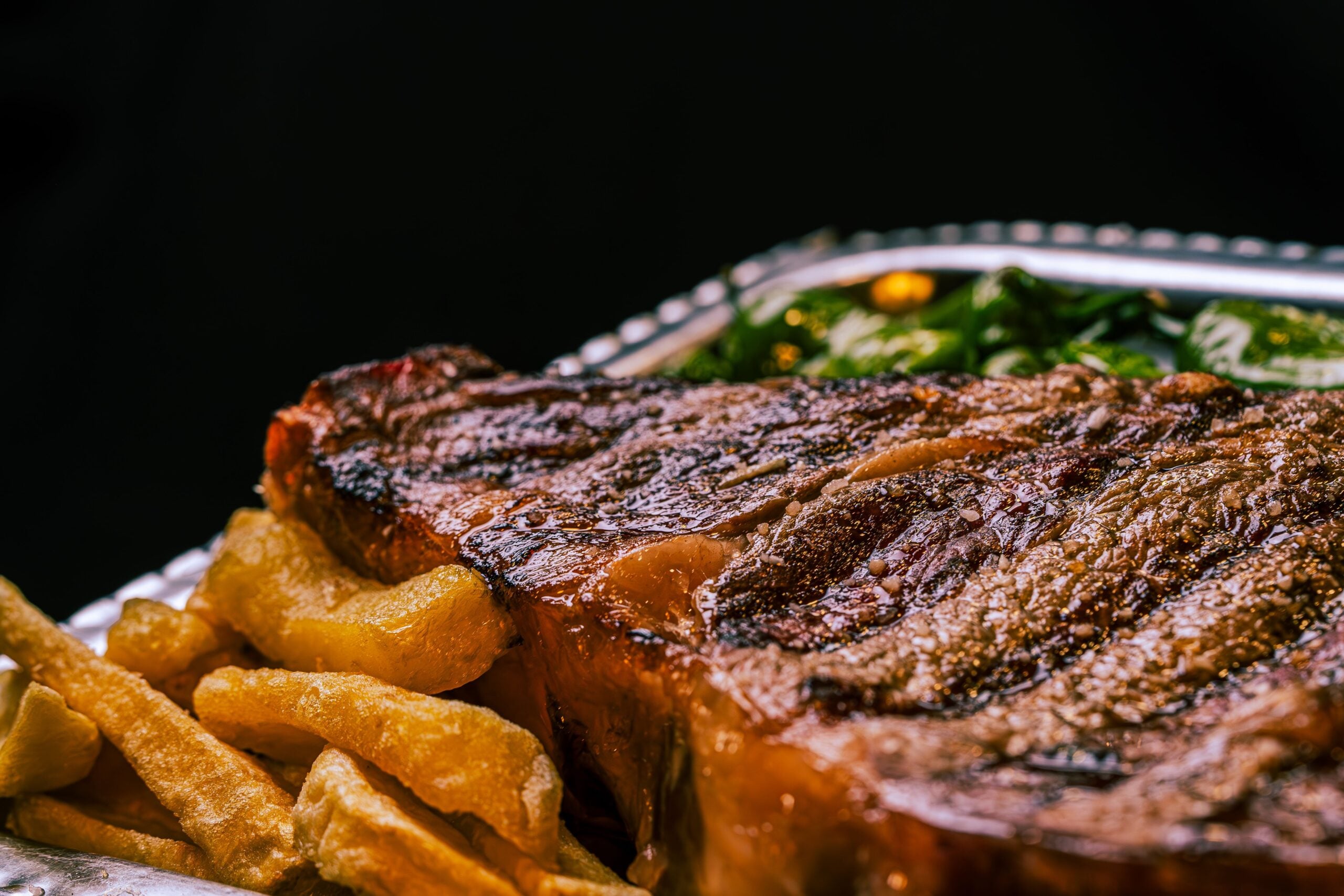 A close-up of a grilled steak served with golden French fries and sautéed greens on a silver plate, set against a black background.