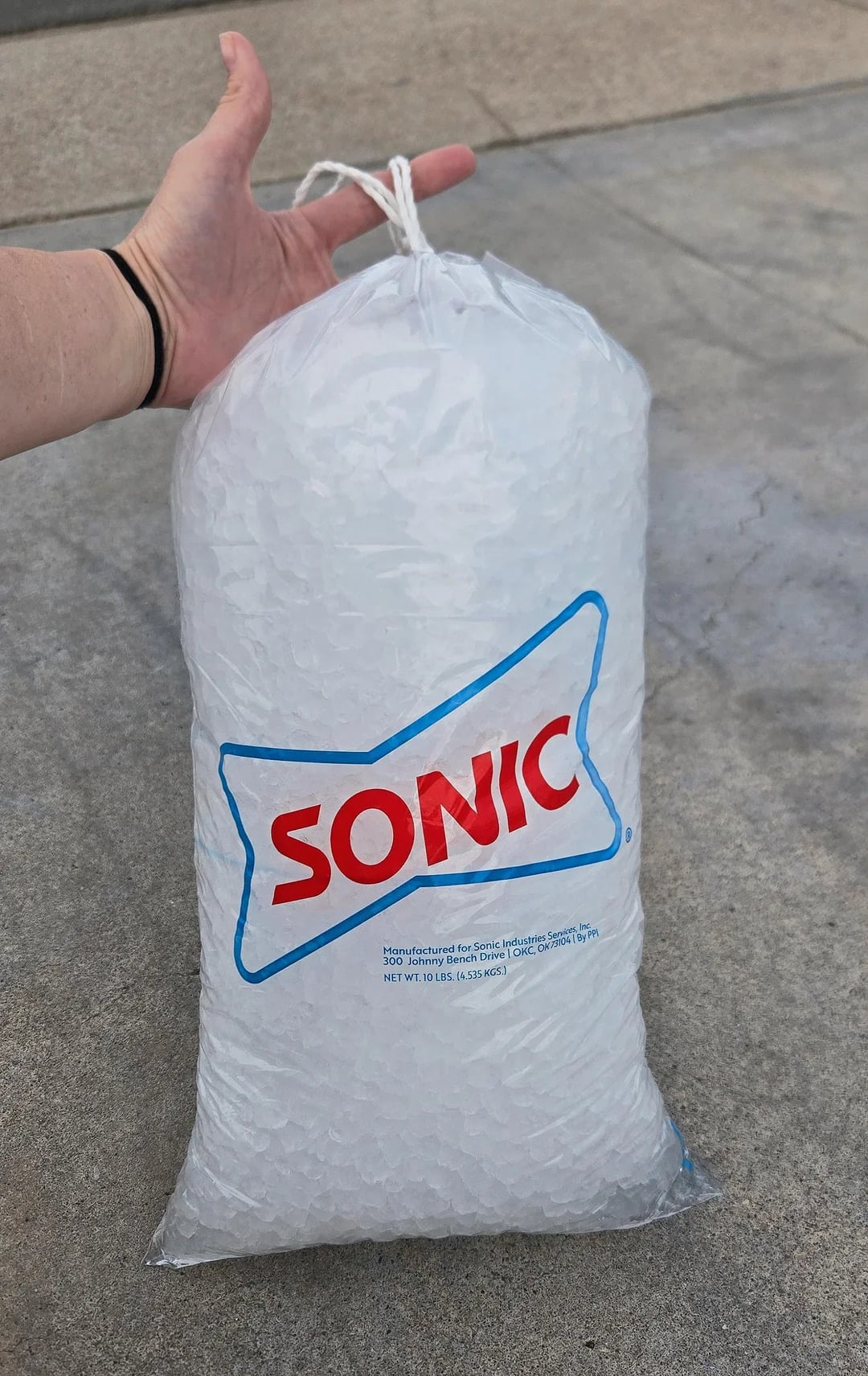 A hand holds a large, clear plastic bag of ice with the Sonic logo printed in red and blue—one of the iconic fast-food grocery store products—resting on a concrete surface.