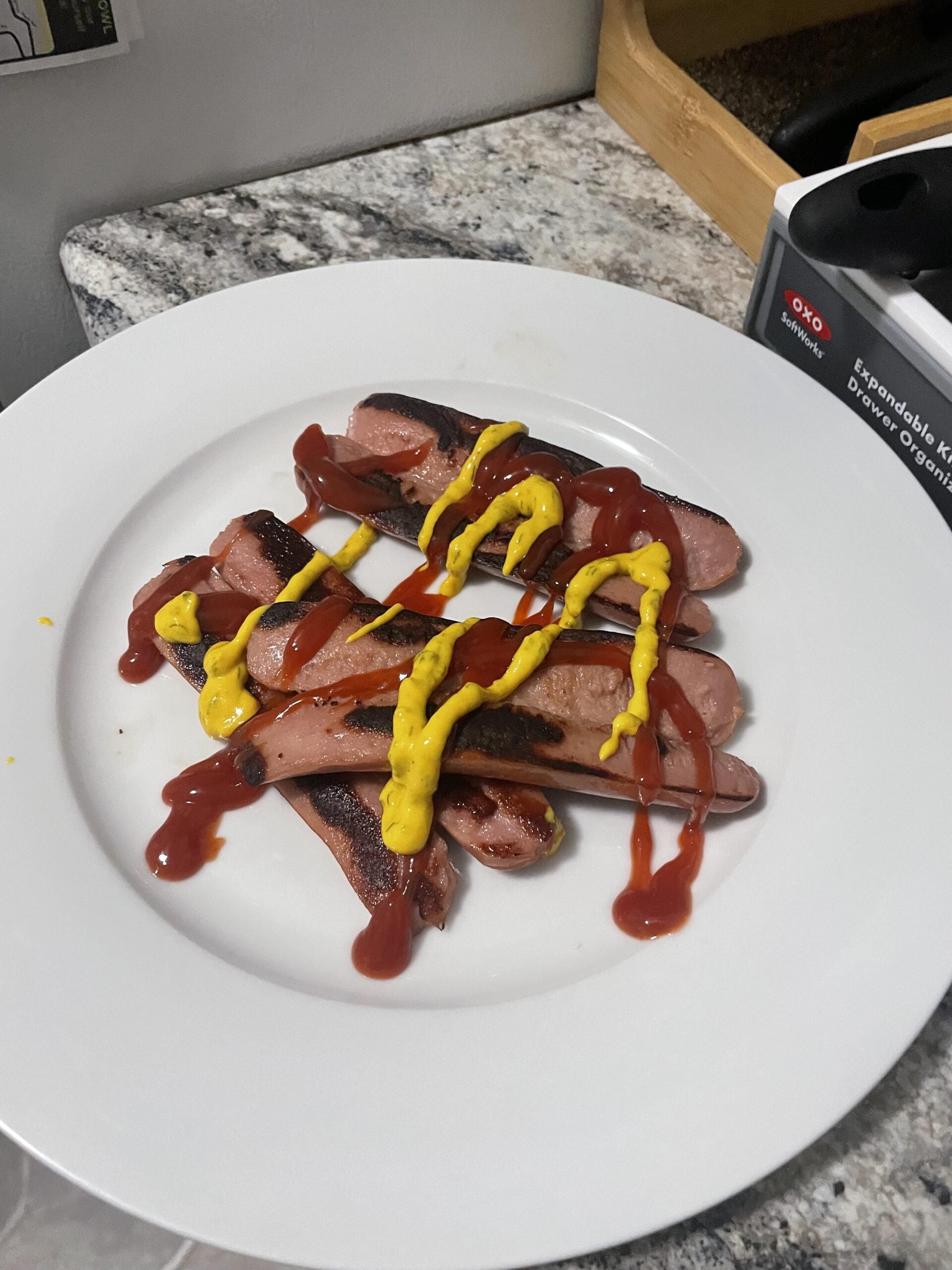 A white plate holds sliced grilled hot dogs topped with wavy lines of ketchup and mustard, sitting on a granite countertop near a black kitchen tool box.