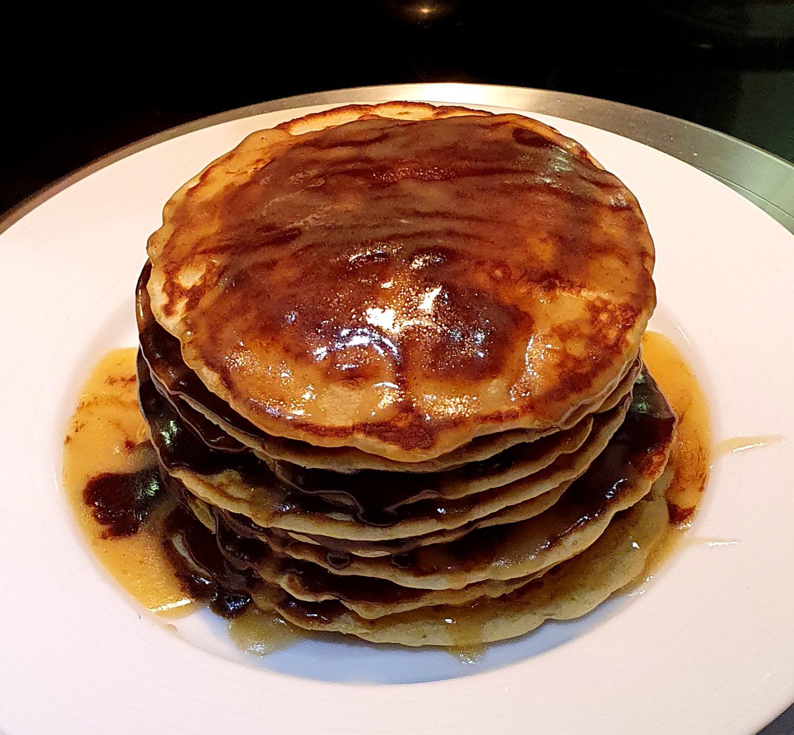 A stack of golden-brown homemade pancakes sits on a white plate, generously covered in syrup that drips down the sides.