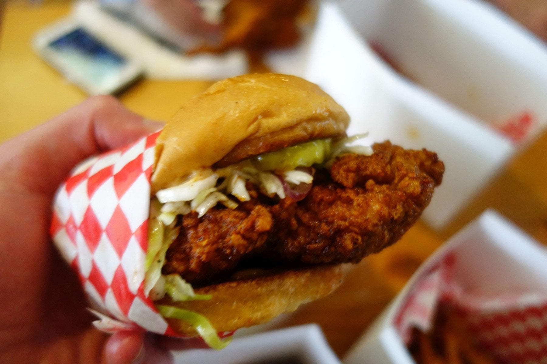 A hand holds a fried chicken sandwich with lettuce and sauce, wrapped in red and white checkered paper. Blurred background shows food containers and a phone on a table.