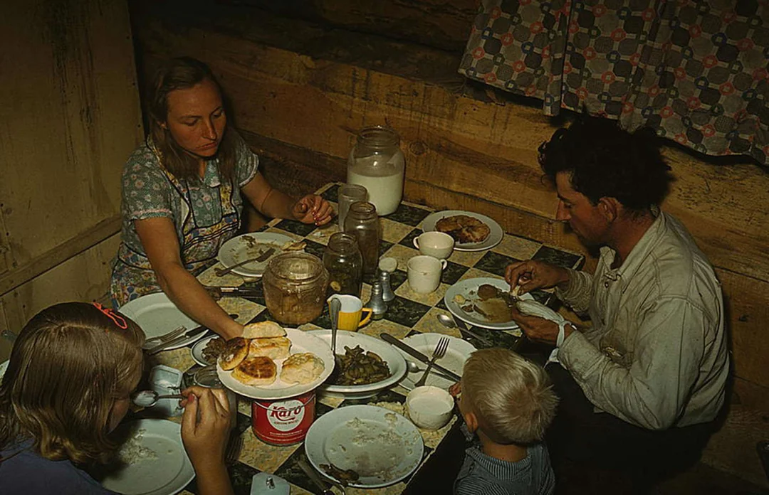 A family of four sits around a small table in a rustic wooden cabin, enjoying nostalgic dinners with biscuits, green beans, and milk, as jars and plates are spread out before them.