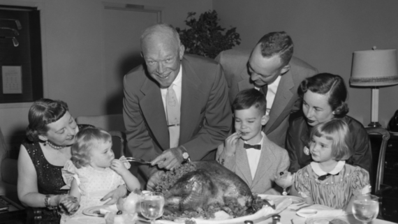 A family gathers around a dining table, smiling as an older man carves a large turkey. Reminiscent of a classic 1950s family dinner, three children and three adults are present, enjoying a festive meal together in a warmly lit room.