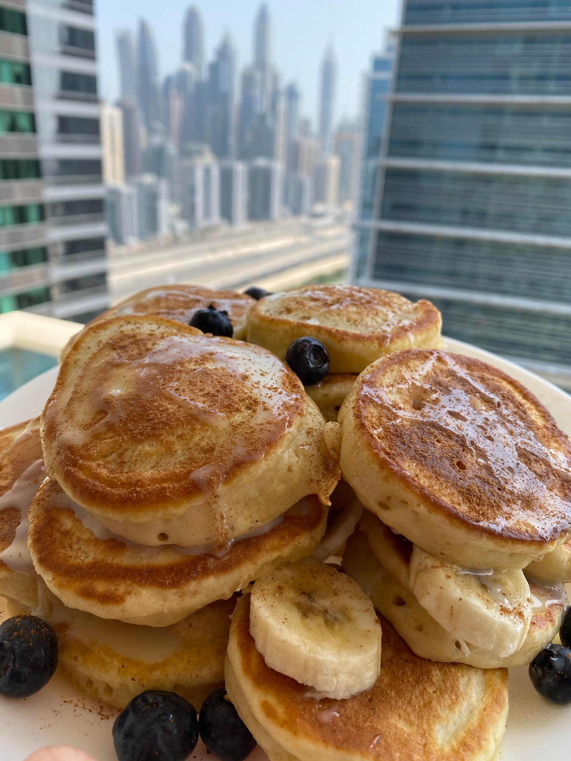 A plate of homemade pancakes topped with banana slices, blueberries, and syrup is set on a balcony with a city skyline and tall buildings in the background.