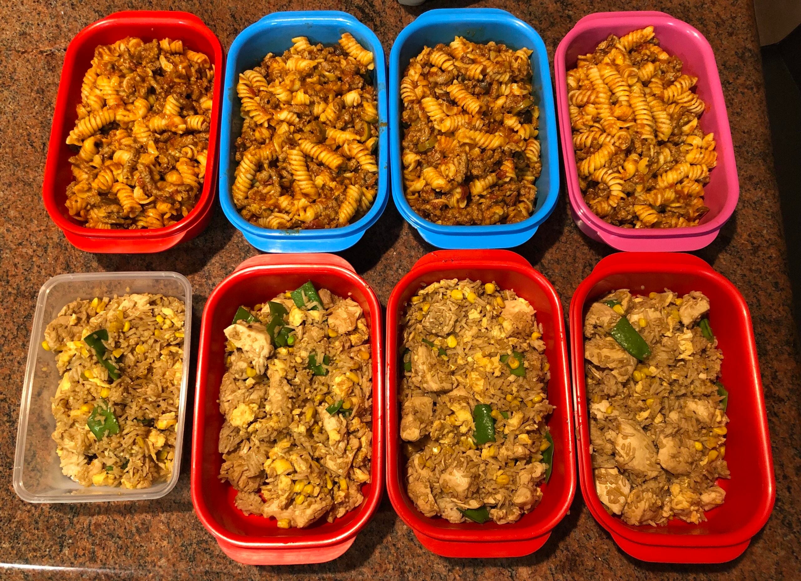 Nine containers with meal prep: the top row has five with twisted pasta and sauce, while the bottom row has four with fried rice, vegetables, and chicken, all arranged on a brown countertop.