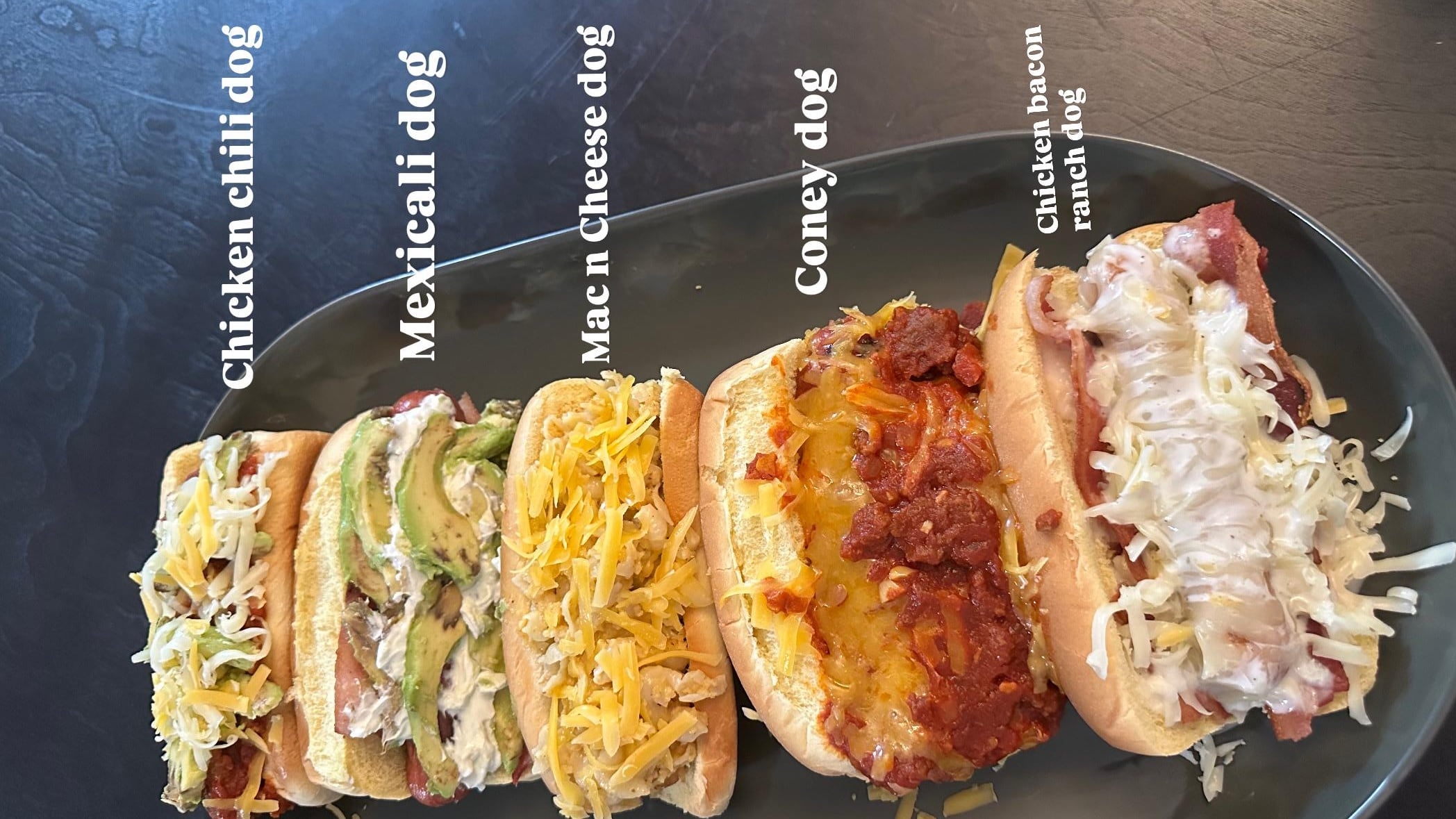 Five unique hot dogs on a black plate, each topped differently: chicken chili, Mexicali with avocado, mac and cheese, Coney-style with chili, and chicken-bacon ranch with shredded cheese. Labels identify each hot dog.