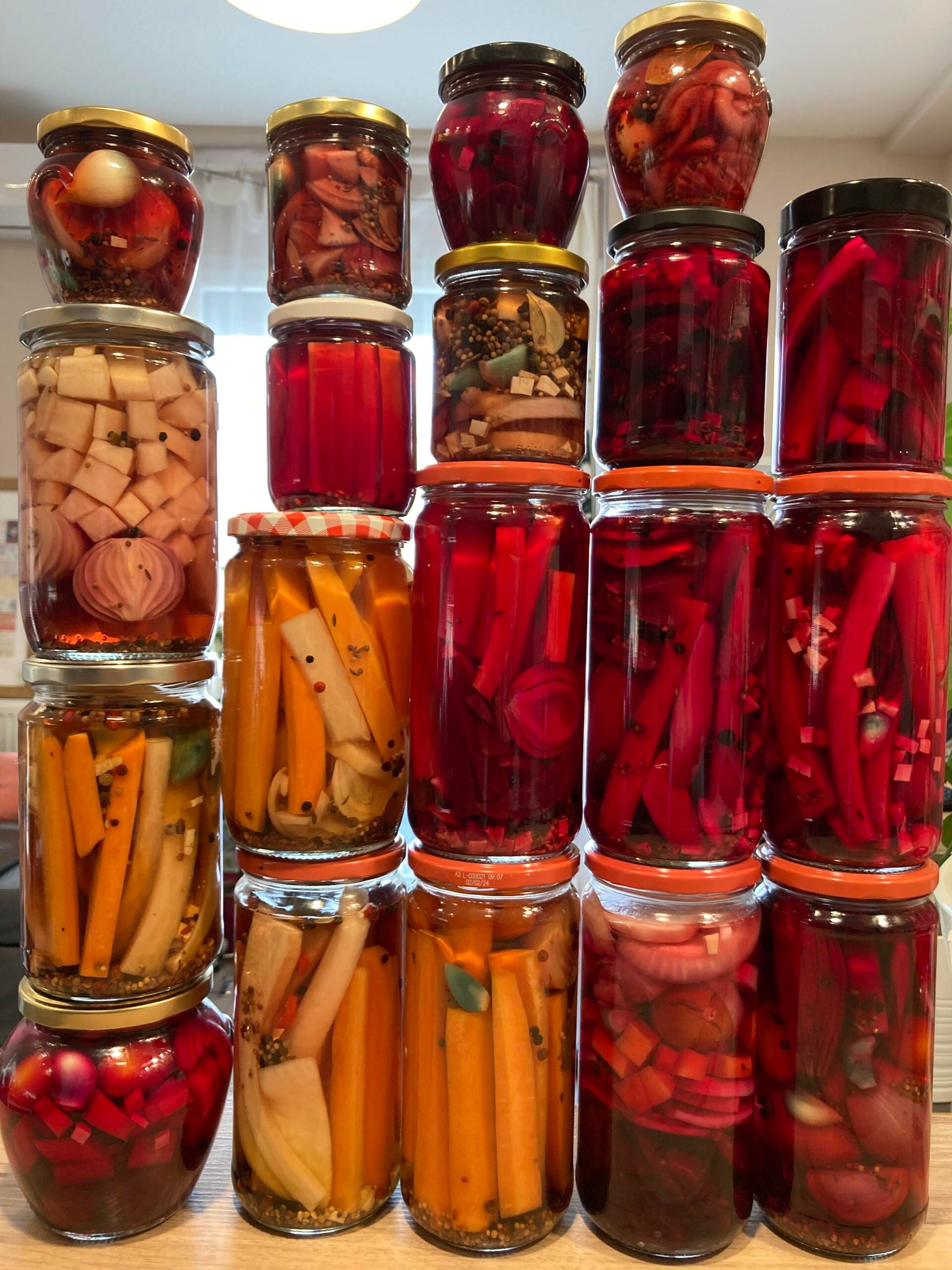 Jars of assorted pickled vegetables, including carrots, radishes, onions, and beets, are stacked in three tall columns on a kitchen counter, showcasing a variety of vibrant colors and shapes.