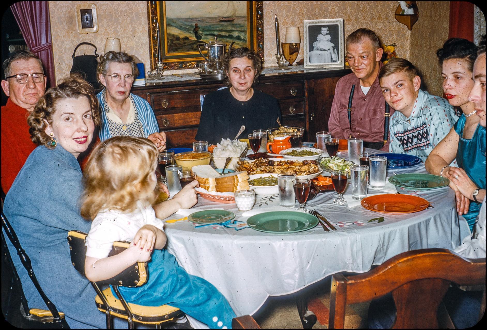 Nine people of various ages sit around a dining table set with food and drinks, smiling and talking in a warmly decorated room, reminiscent of nostalgic dinners, with a painting and lamp on a sideboard in the background.