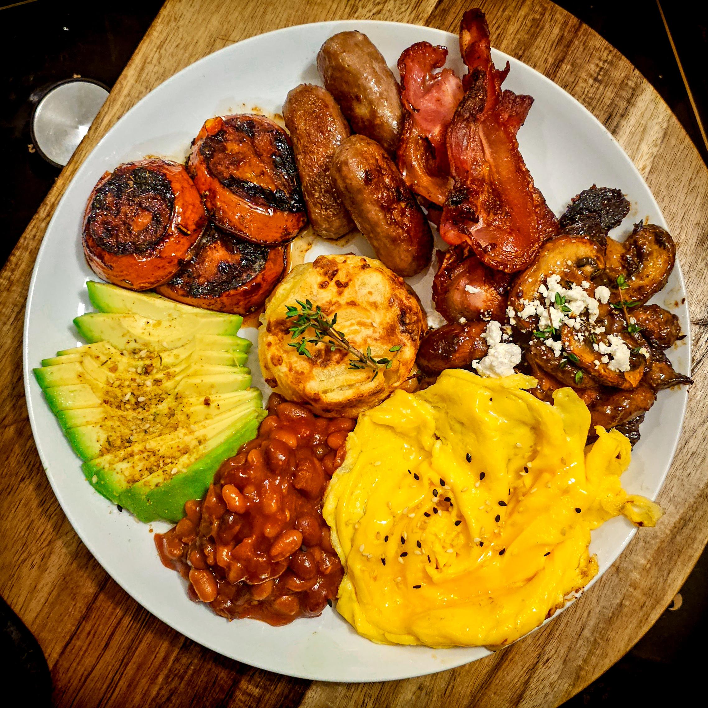 A breakfast plate with scrambled eggs, avocado slices, baked beans, sweet potato, sausage, bacon, grilled mushrooms with feta, and a biscuit, served on a wooden board.