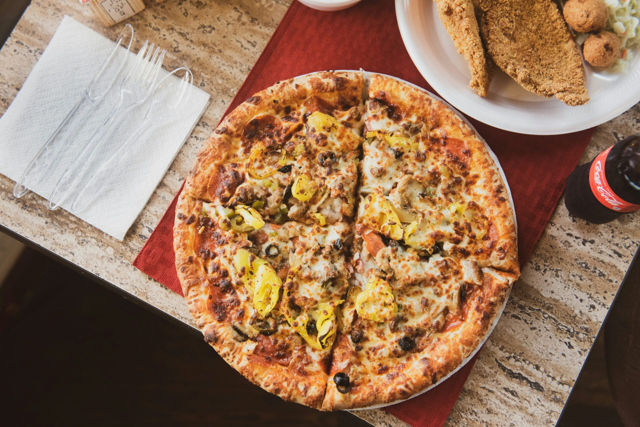 A large pizza topped with cheese, sausage, olives, and banana peppers on a table next to fried food, a bottle of Coke, and plastic cutlery on a napkin.