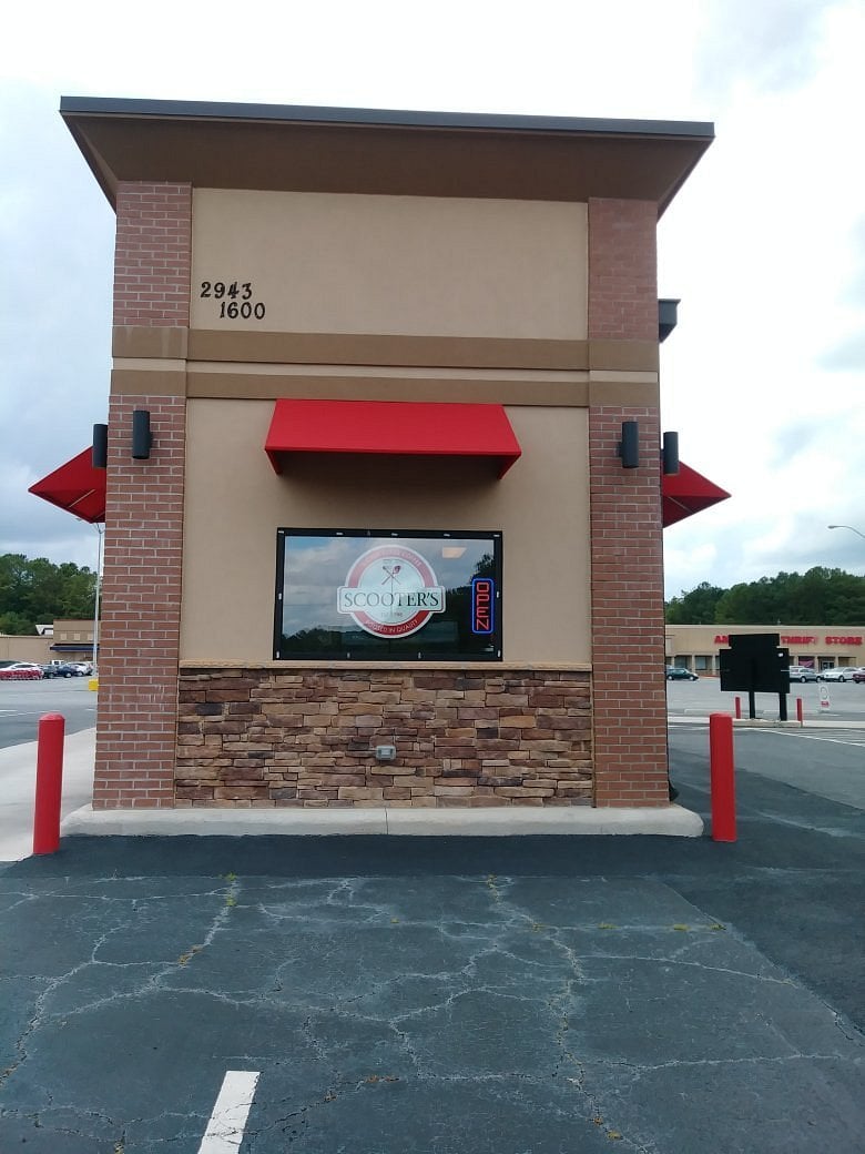 A building with a red awning, sign for "Scooters" below a window, and address numbers "2943" and "1600" above it. There are red posts and a parking lot in front, reminiscent of many regional fast-food chains, with stores in the background.