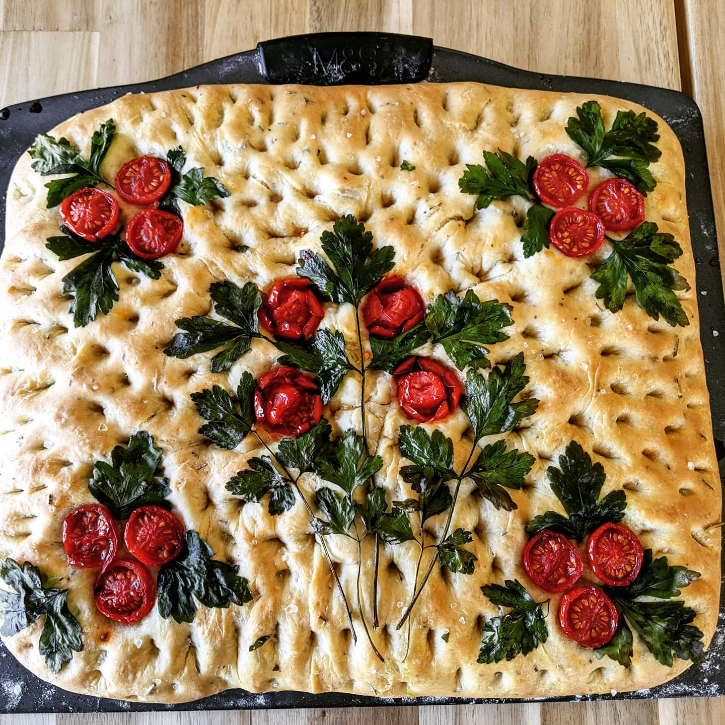 A rectangular focaccia bread decorated with clusters of cherry tomato slices and parsley leaves, arranged to resemble flowers and stems. The bread is golden brown and dotted with dimples.