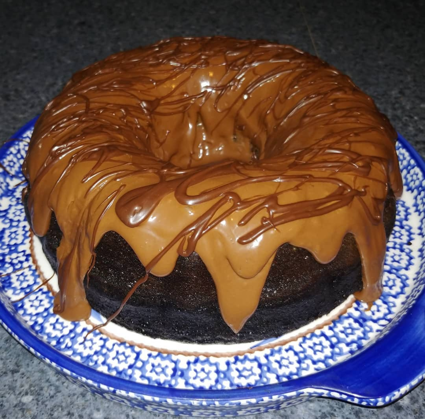 A homemade dessert, this chocolate bundt cake is topped with a thick layer of caramel and drizzled with chocolate, beautifully displayed on a decorative blue and white plate.