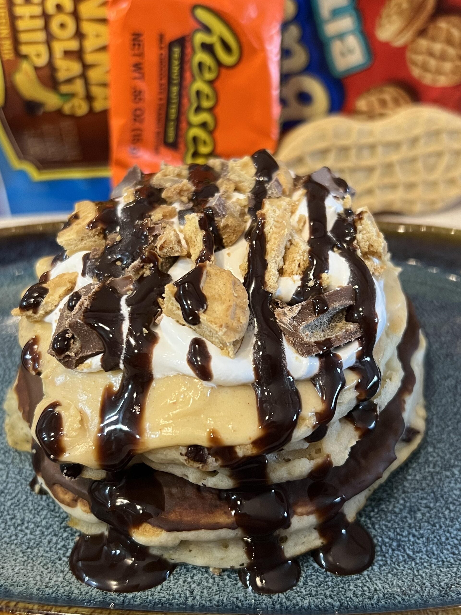 A stack of pancakes topped with whipped cream, crumbled Reese’s candy, chocolate syrup, and peanut butter, with candy packages in the background—a decadent treat perfect for fans of homemade desserts.