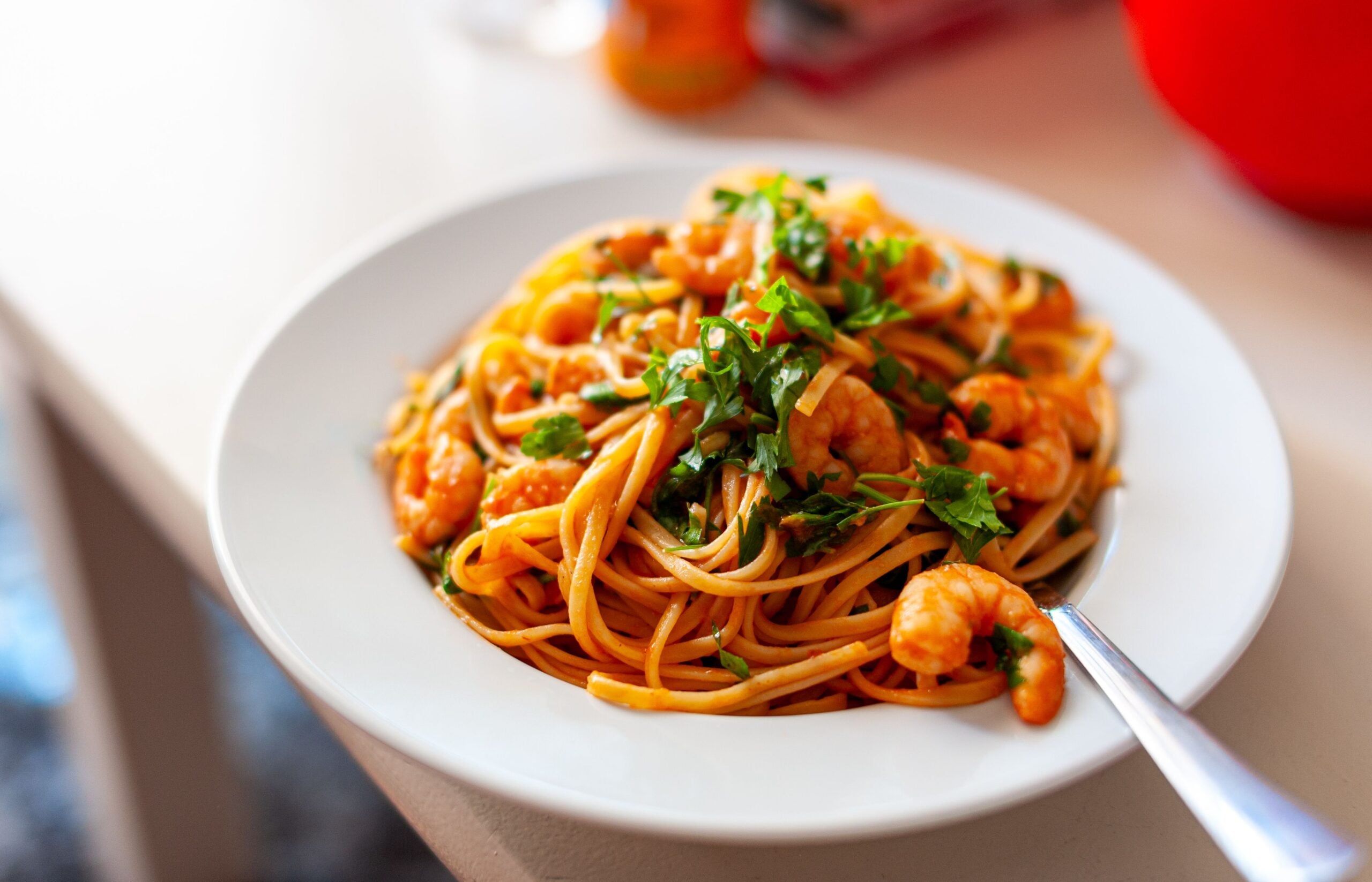 A white plate filled with spaghetti, shrimp, and fresh herbs sits on a table. A fork rests on the plate, ready to be used. The dish is brightly lit, highlighting the colors and textures.
