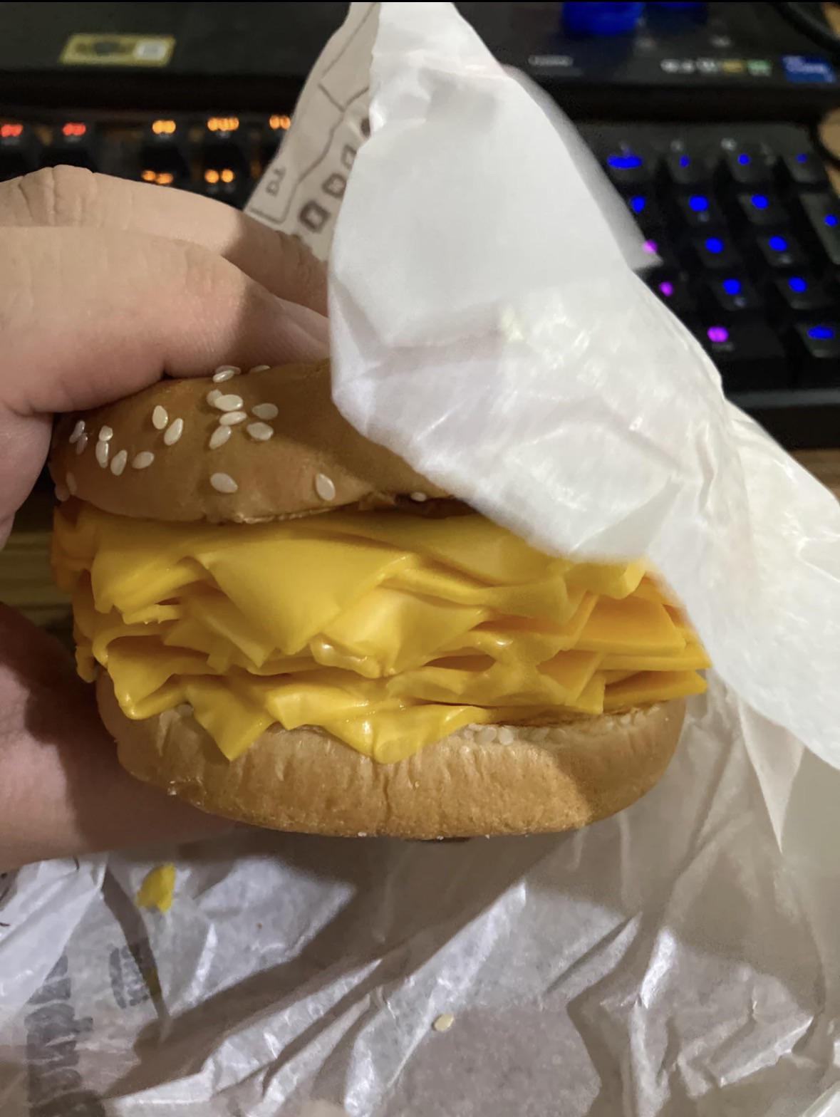 A hand holds a sesame seed bun filled with an excessive amount of folded American cheese slices, partially wrapped in white paper. A keyboard is visible in the background.