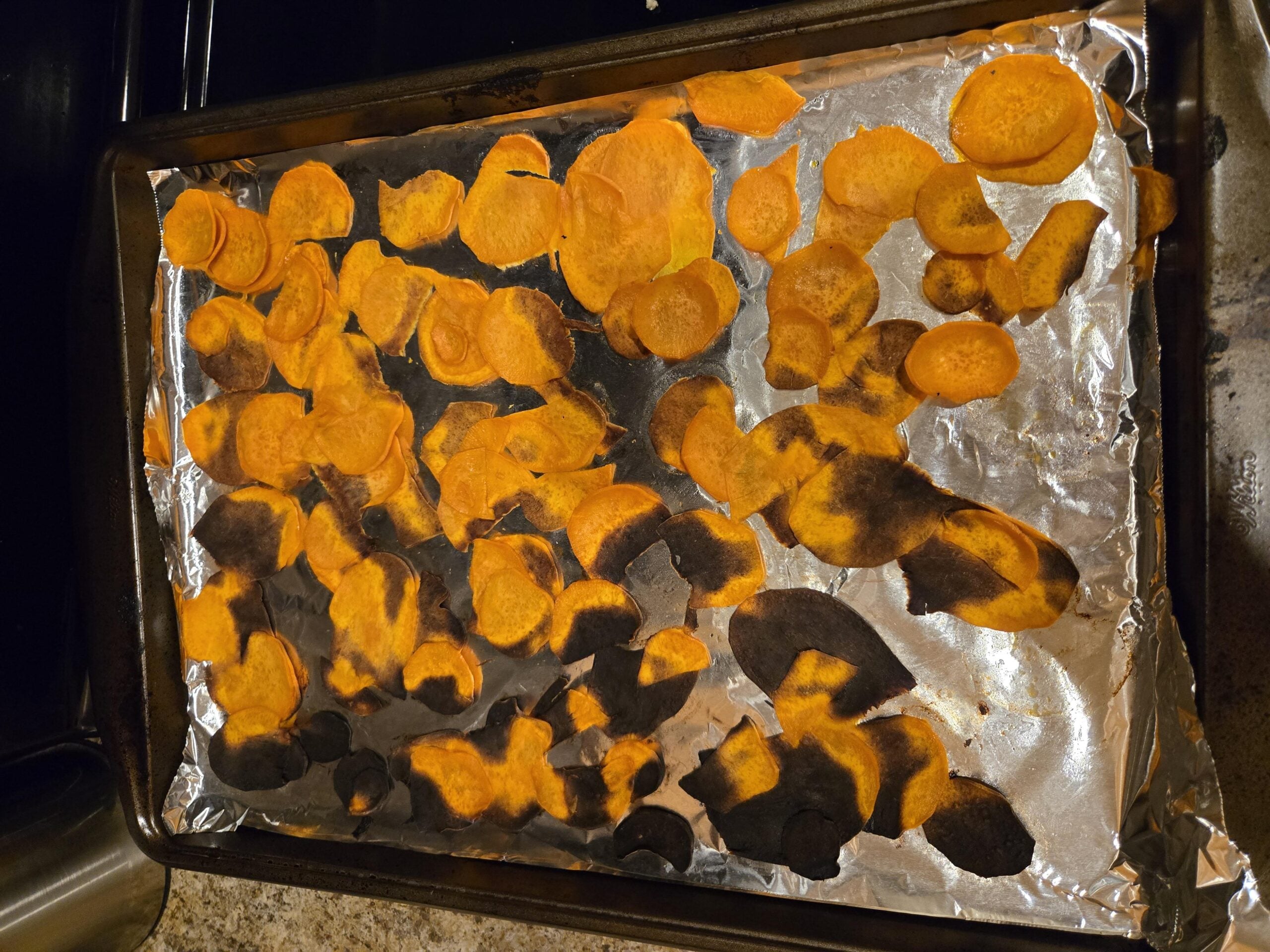 A baking sheet covered with aluminum foil holds thinly sliced sweet potato chips, some of which are burnt and blackened while others appear properly cooked—a classic scene straight out of hilarious cooking disasters. The sheet is on a countertop.