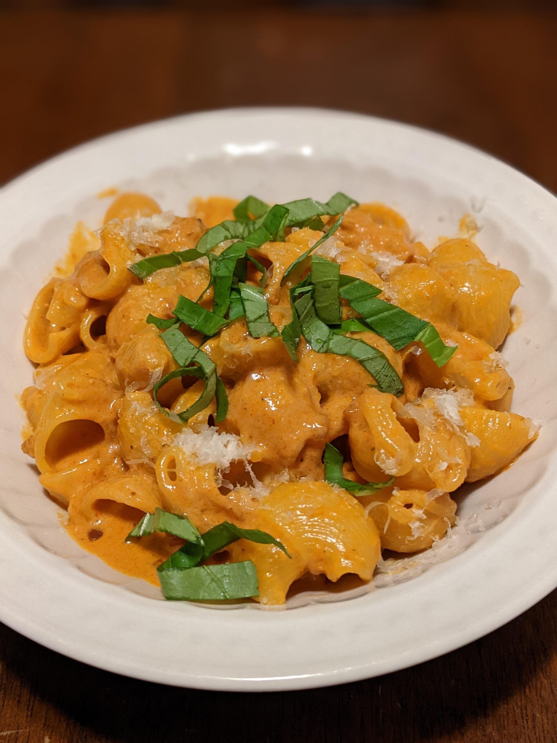 A white bowl filled with creamy pasta topped with shredded cheese and fresh basil ribbons, set on a wooden table.