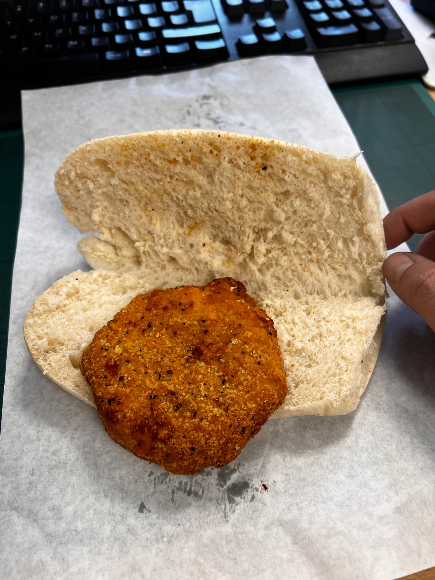 A hand opens a bun with a single breaded chicken patty inside, resting on white paper with a keyboard visible in the background.