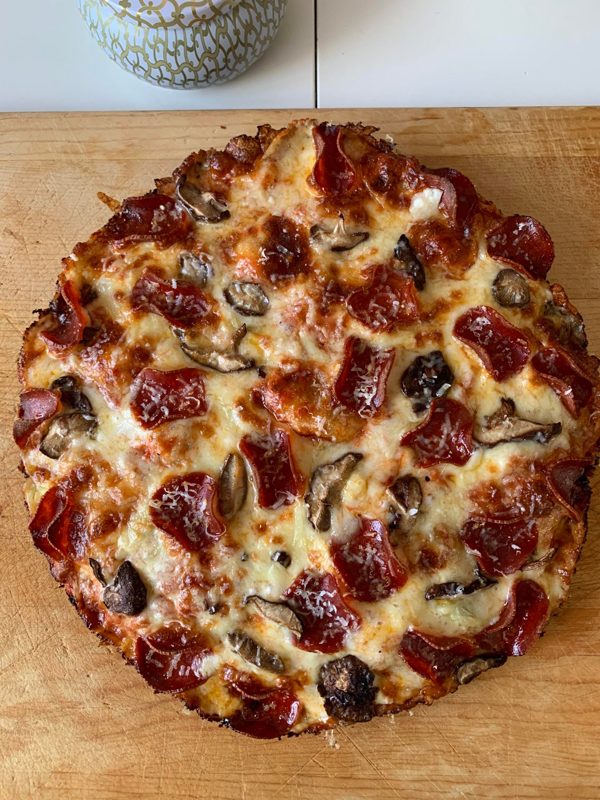 A homemade pizza topped with pepperoni, mushrooms, and melted cheese sits on a wooden cutting board. A patterned container is visible in the background.