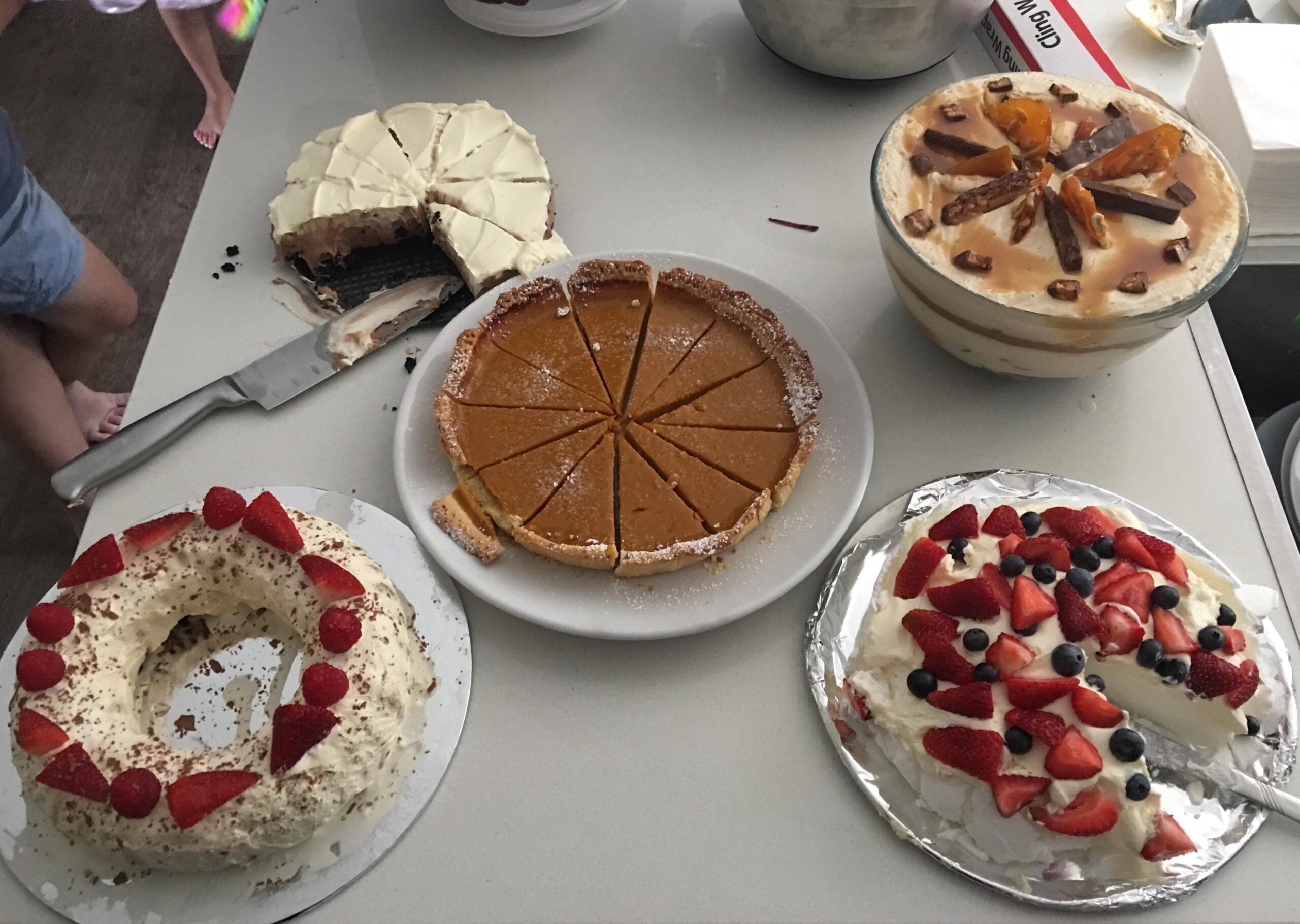 Four homemade desserts on a table: a sliced cheesecake, a whole pumpkin pie, a glass trifle with toffee bits, and two cakes topped with cream and berries. A person stands nearby, partly visible.