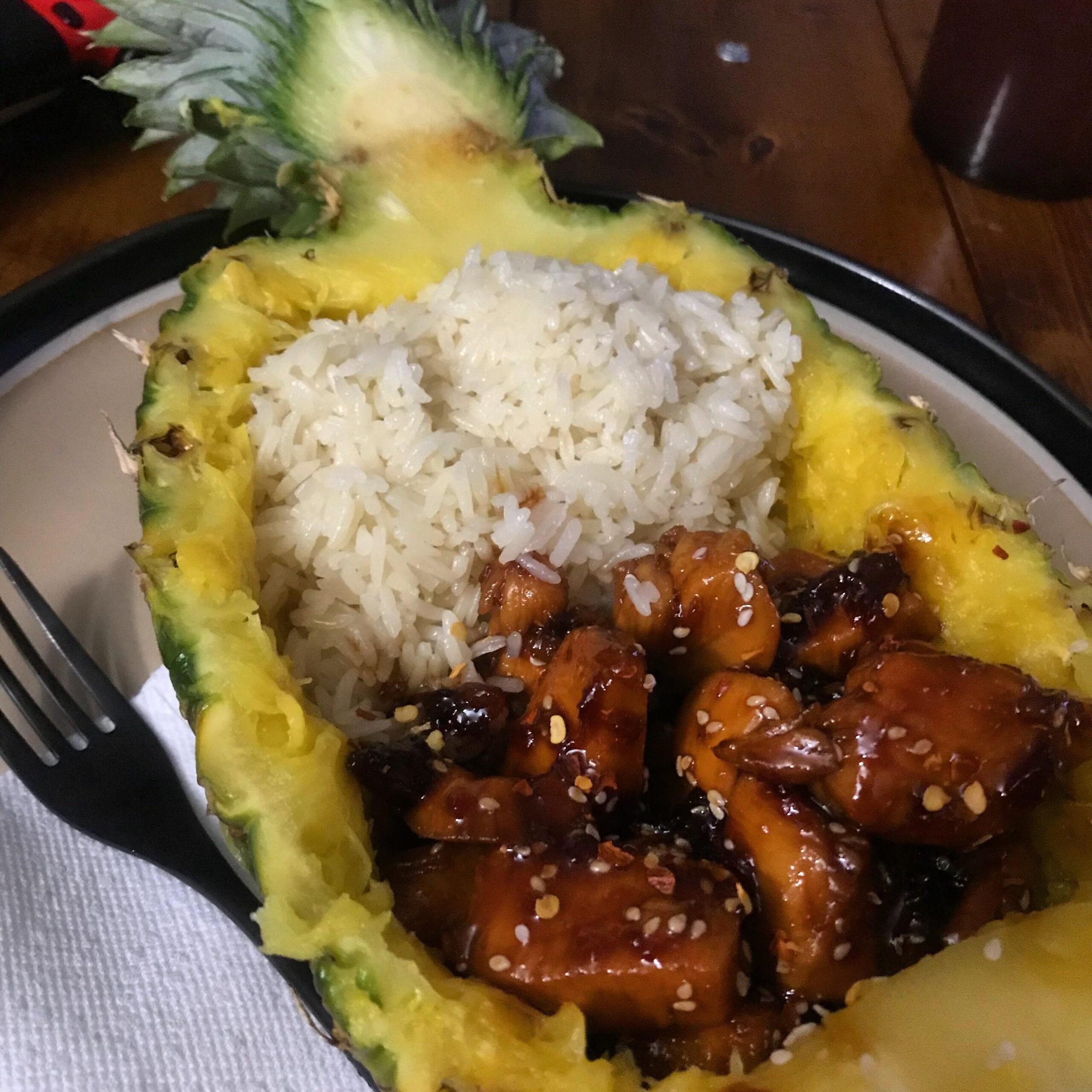 Half a pineapple used as a bowl, filled with white rice and glazed chicken pieces topped with sesame seeds. A fork and napkin are placed beside the pineapple on a plate.