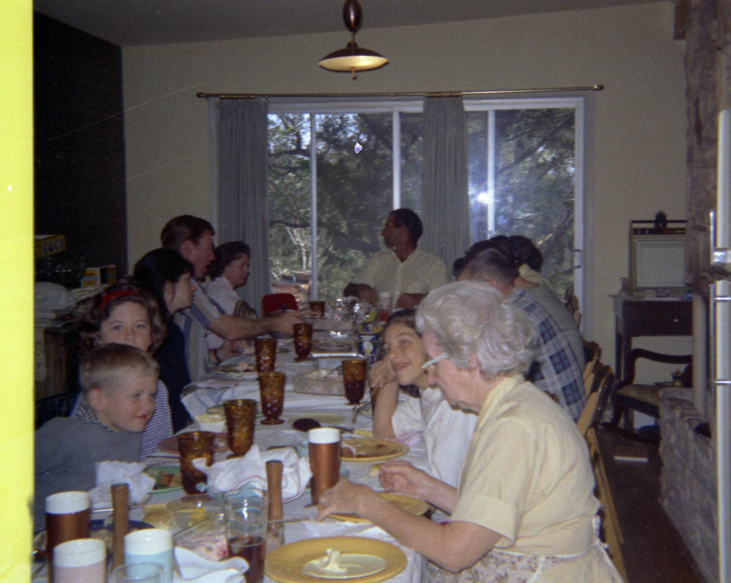 A family of various ages sits around a dining table set with food and drinks, enjoying nostalgic dinners together in a home with large windows and a stone fireplace.