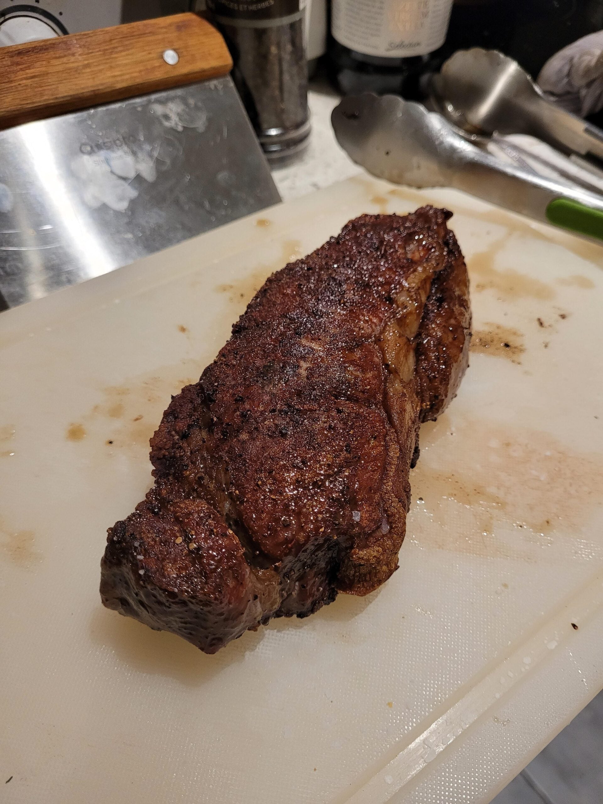 A cooked, seasoned steak rests on a white cutting board with kitchen utensils and a bottle visible in the background.