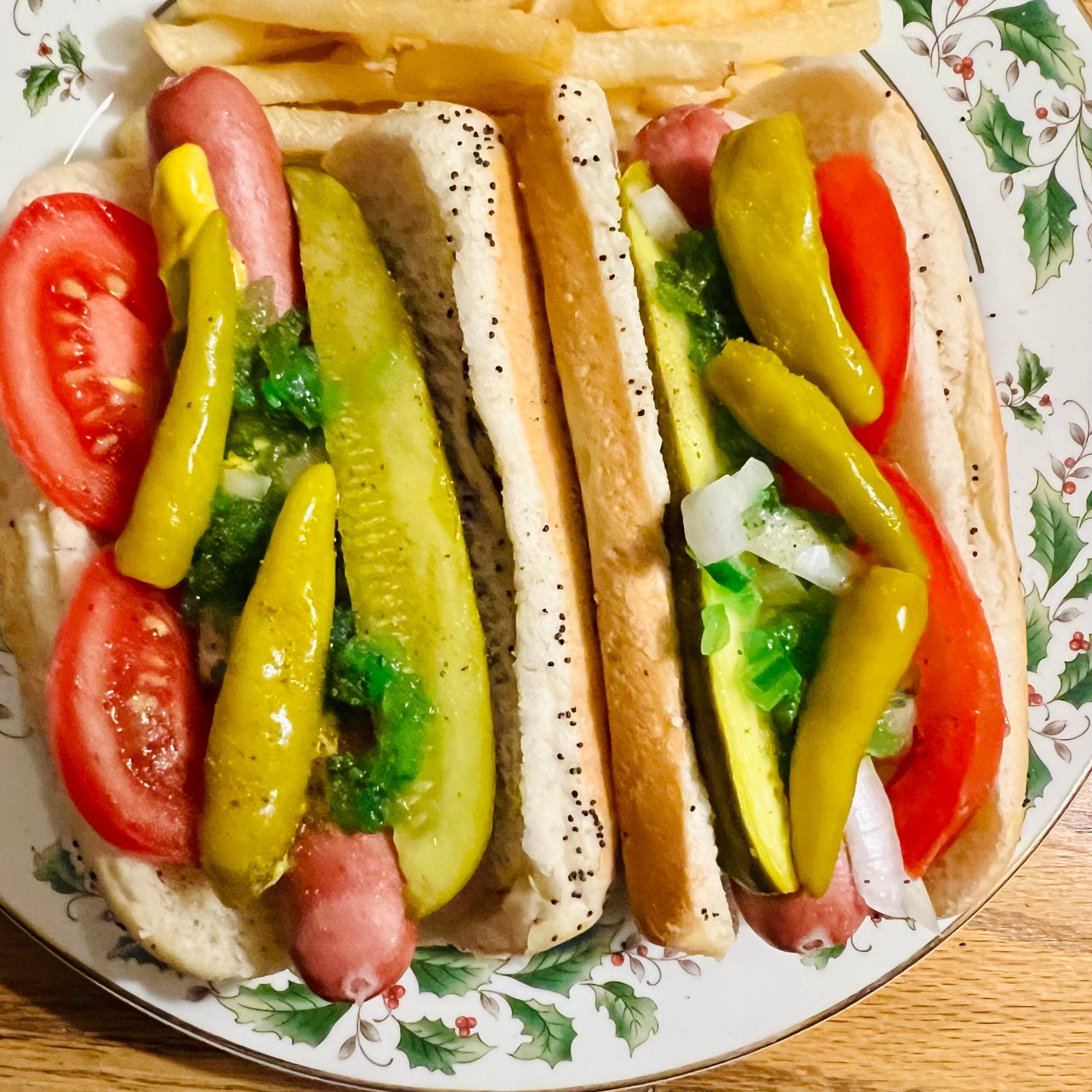 Two Chicago-style hot dogs in buns with pickles, tomatoes, onions, sport peppers, relish, and mustard on a festive plate, served with French fries.