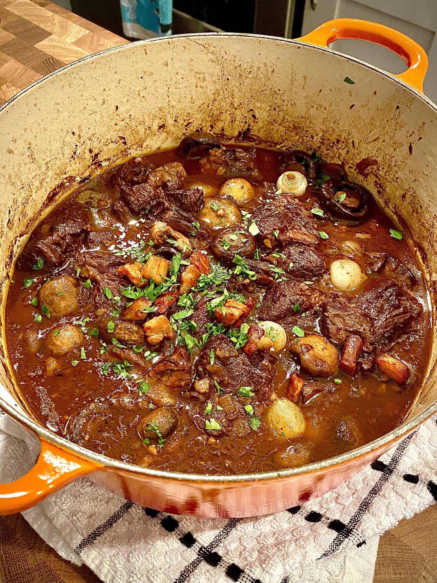 A pot of beef stew with pieces of meat, carrots, onions, and herbs in rich brown sauce, served in an orange Dutch oven on a checkered towel.