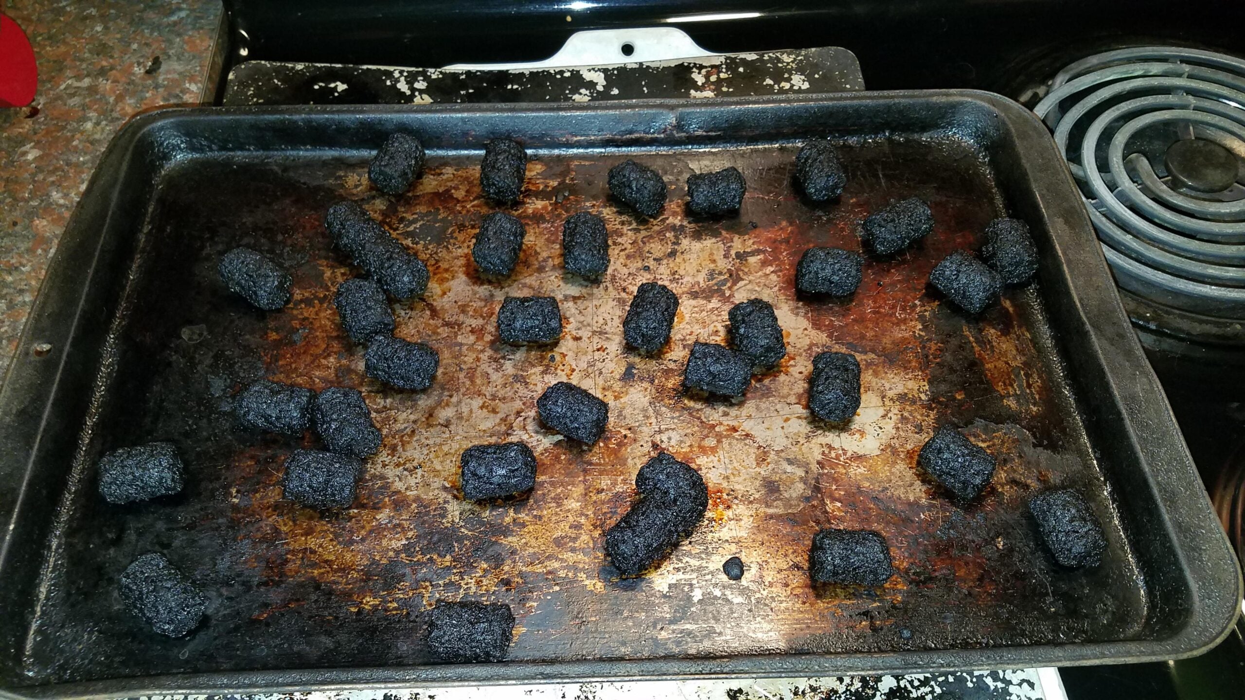 A baking sheet with several severely burnt tater tots scattered across it, all blackened and charred beyond recognition.