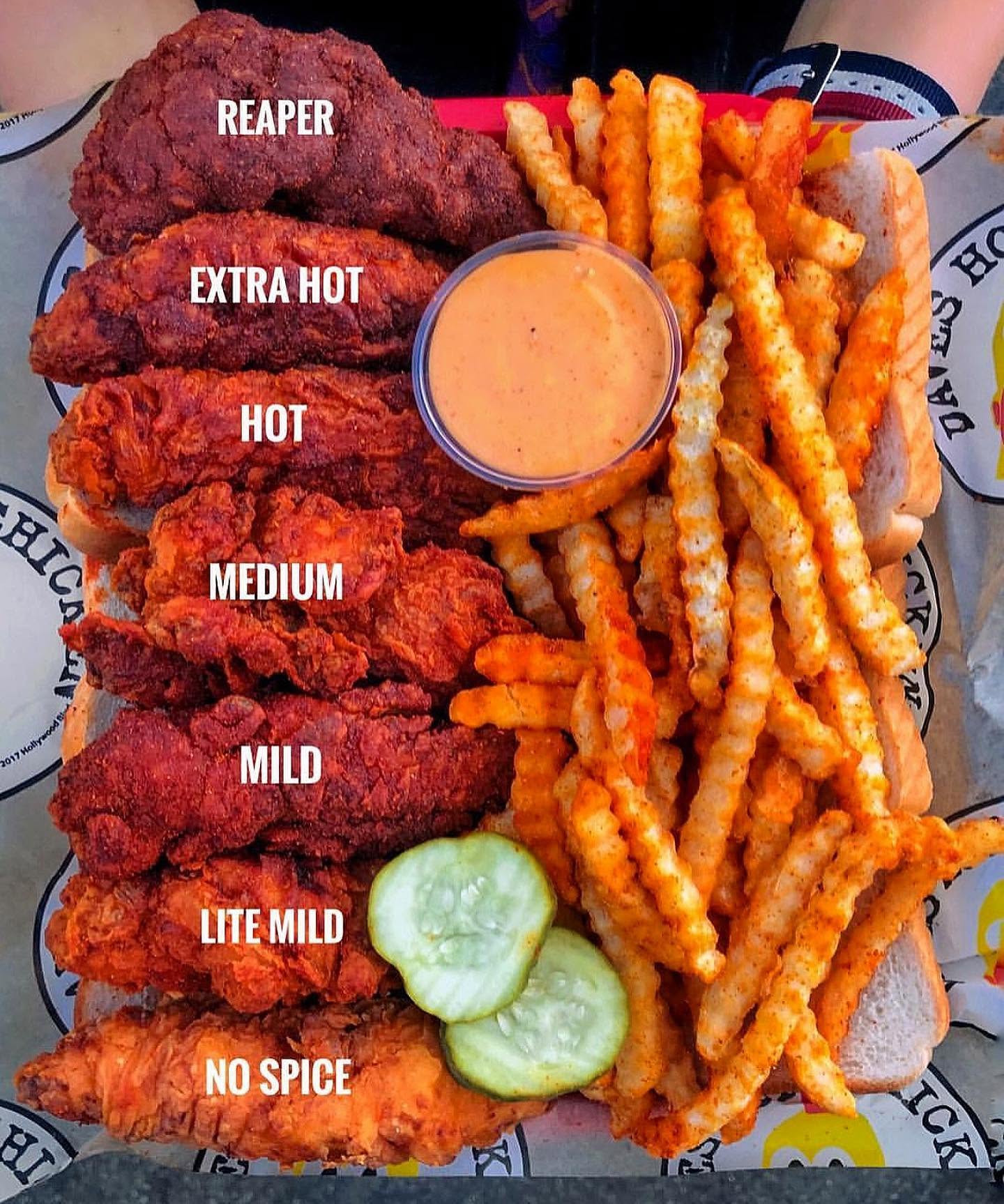 A tray of chicken tenders, reminiscent of top fried chicken chains, is labeled by spice level from "No Spice" to "Reaper," with crinkle-cut fries, pickles, toast, and a cup of dipping sauce. The spiciness increases from bottom to top.