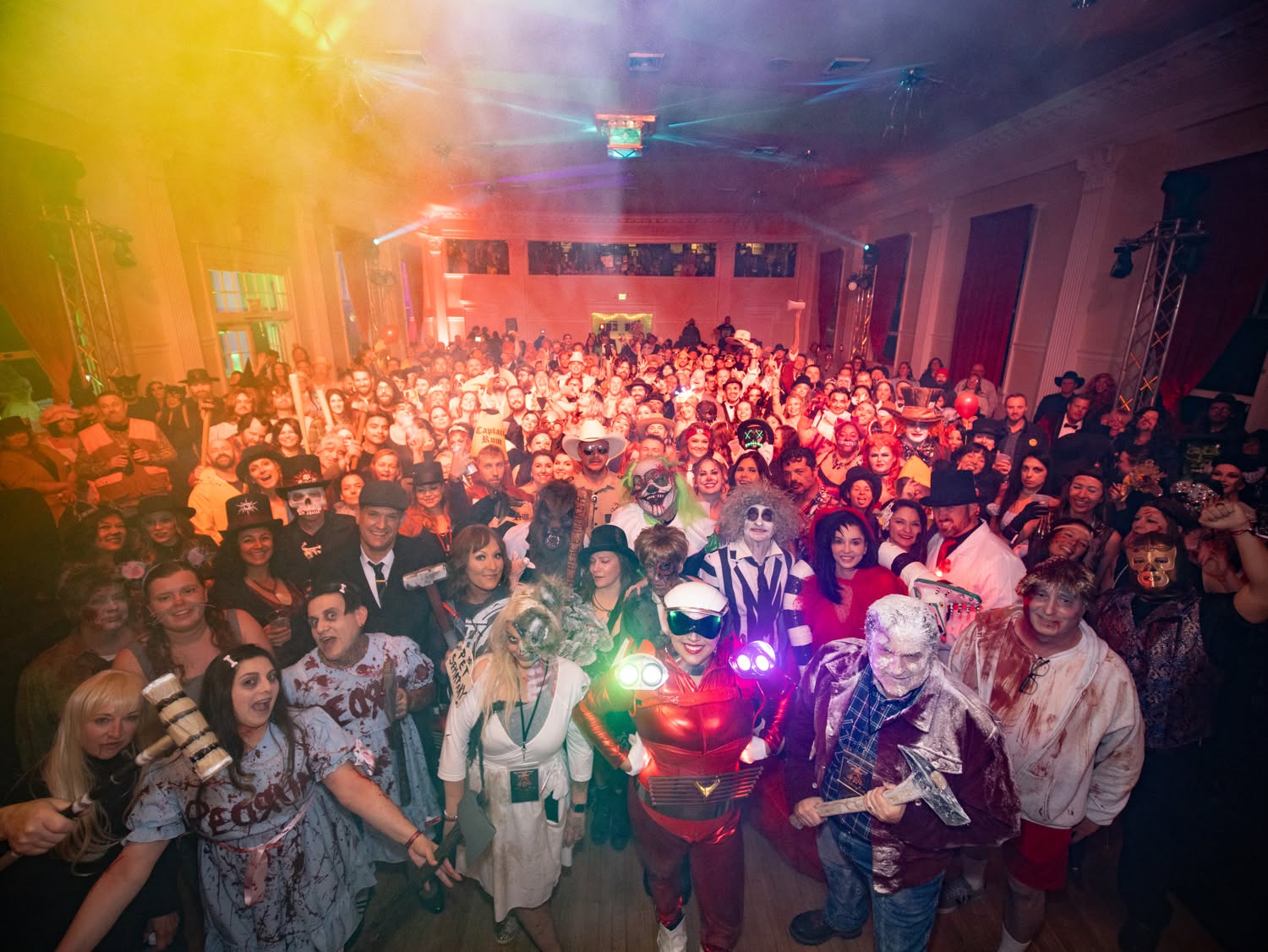 A large group of people in elaborate costumes, including superheroes, zombies, and other spooky Halloween characters, pose together at a lively, colorful indoor event with dramatic lighting and fog effects.