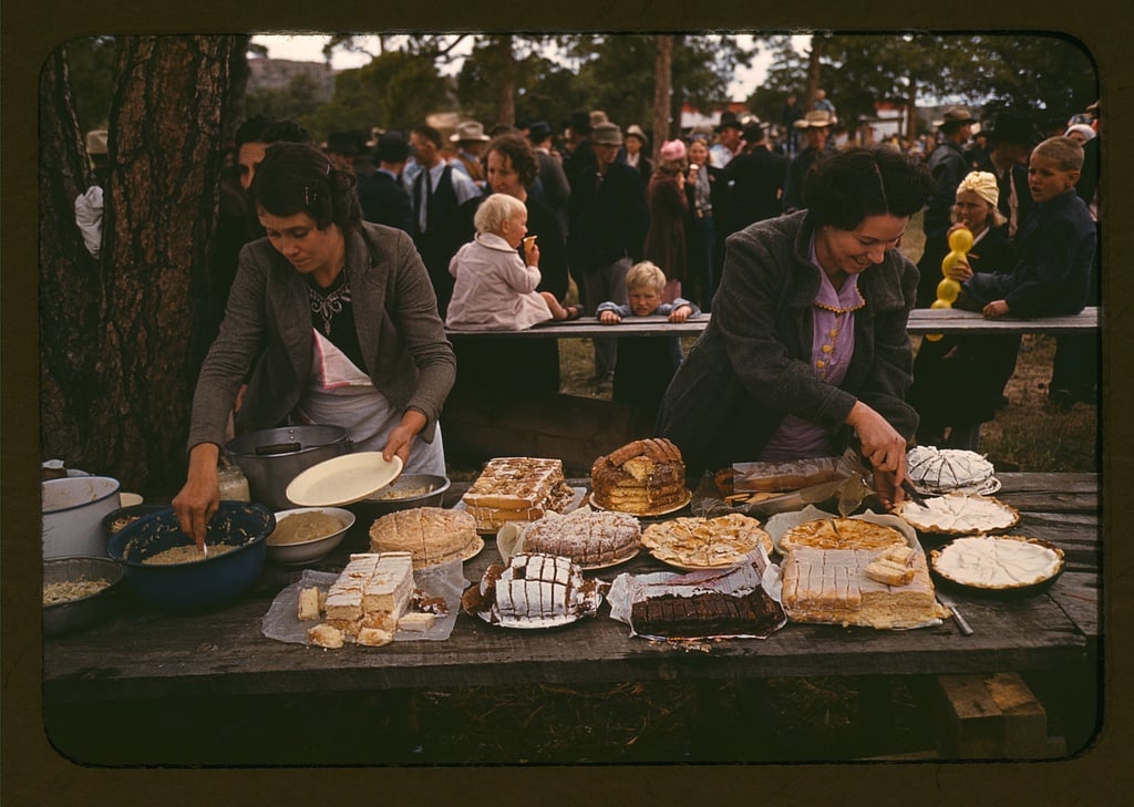 Two women serve a variety of cakes and desserts on a wooden table at an outdoor event, evoking the charm of nostalgic dinners, with a crowd of people gathered in the background.