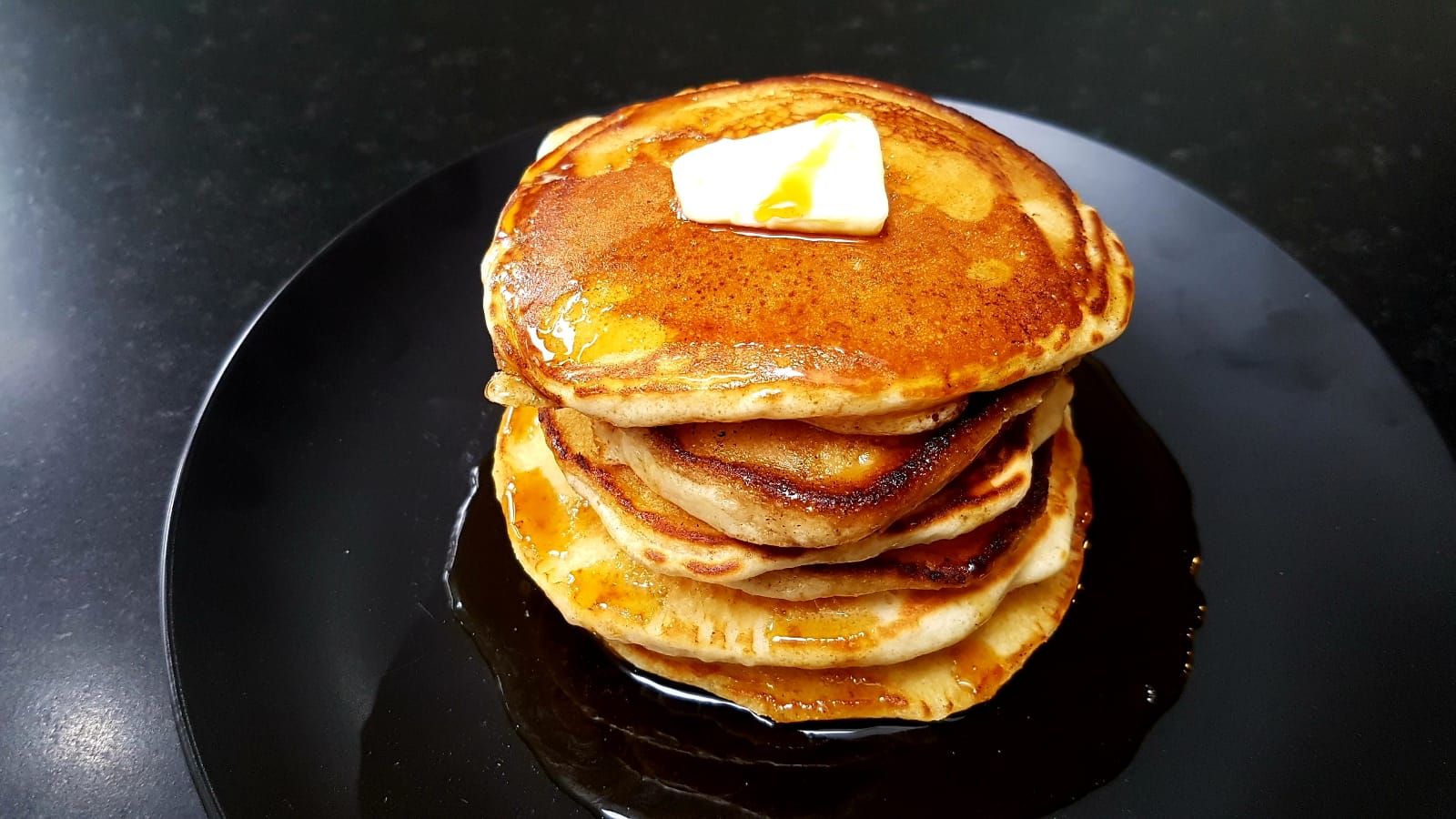 A stack of golden-brown pancakes sits on a black plate, topped with a melting pat of butter and drenched in syrup that pools at the base—a cozy favorite among homemade desserts.