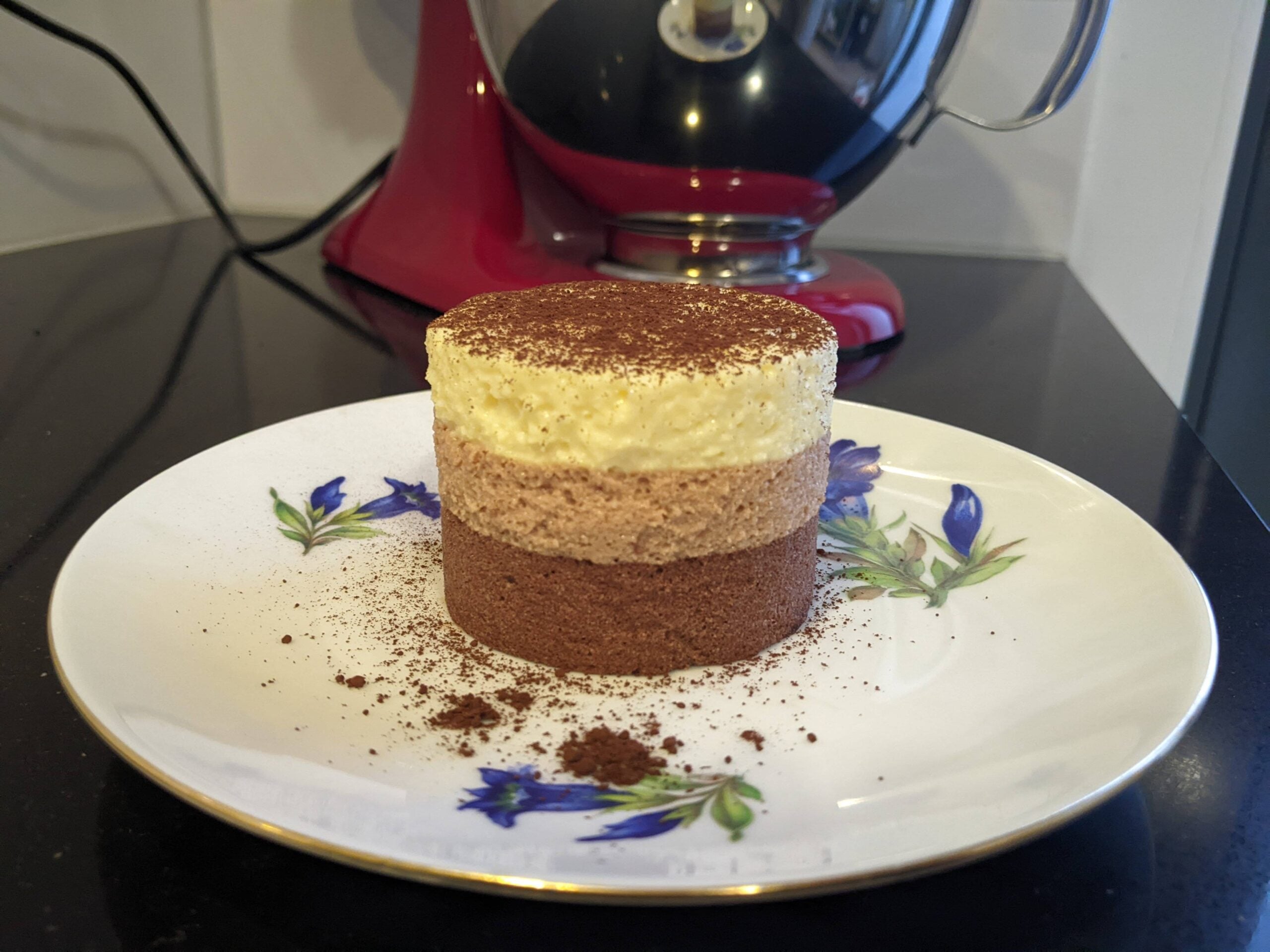 A three-layer mousse cake—chocolate, mocha, and vanilla—sits on a floral plate, topped with cocoa powder. Perfect for lovers of homemade desserts, this decadent treat is displayed on a black countertop with a red stand mixer in the background.