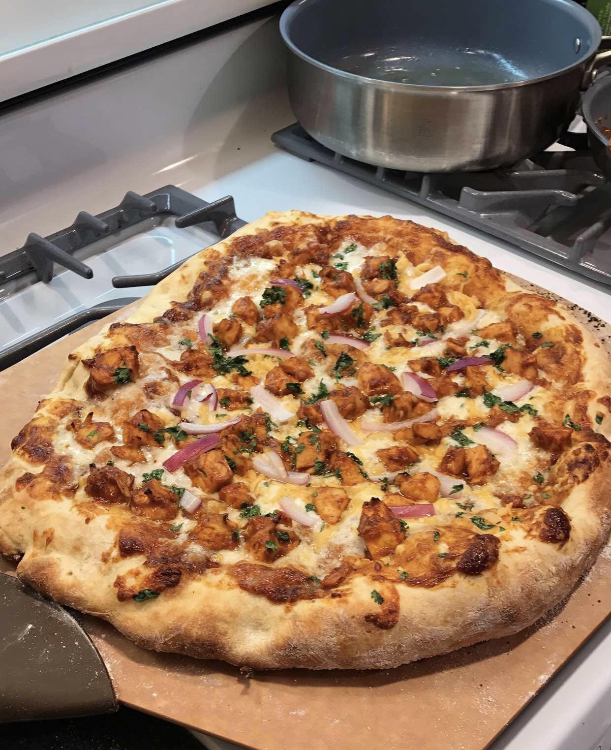 A freshly baked homemade pizza topped with chunks of chicken, red onion slices, cheese, and herbs sits on a baking tray next to a stove with pots and pans.