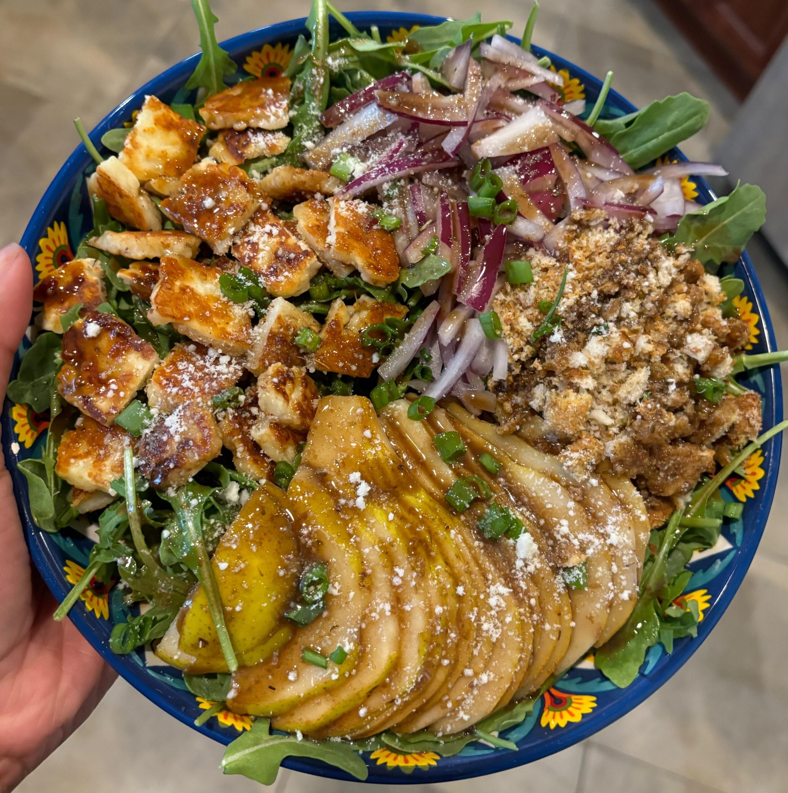 A colorful salad bowl with arugula, grilled halloumi, sliced pears with dressing, red onions, and a crumbly topping, all garnished with herbs and cheese.