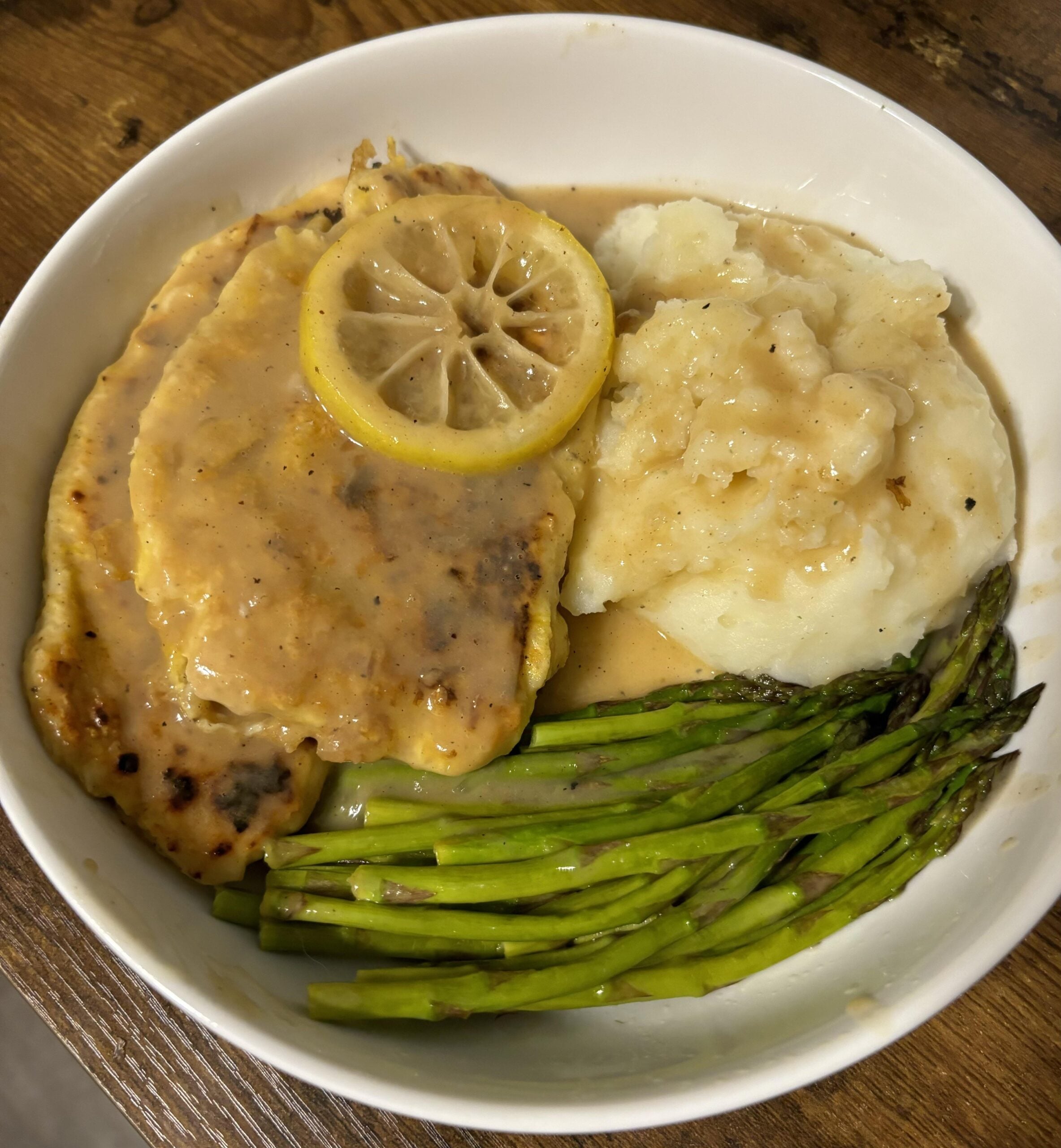 A bowl with lemon chicken in sauce, mashed potatoes with gravy, and steamed asparagus spears on a wooden table.