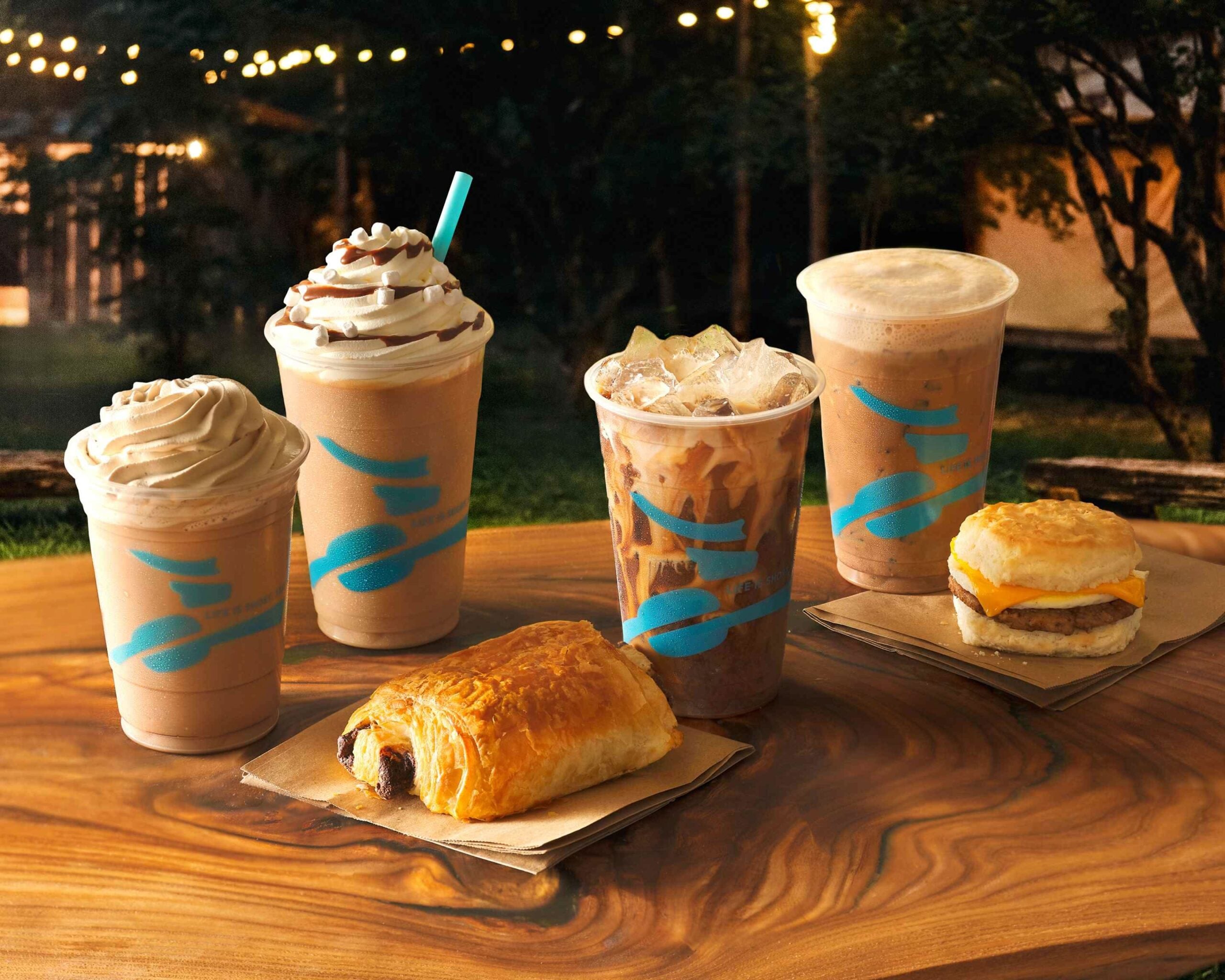 Five drinks and two breakfast foods on a wooden table evoke the cozy vibe of coffee houses: three blended iced coffees, one iced coffee, one hot coffee, a sausage biscuit sandwich, and a chocolate-filled pastry in an outdoor, warmly lit setting.