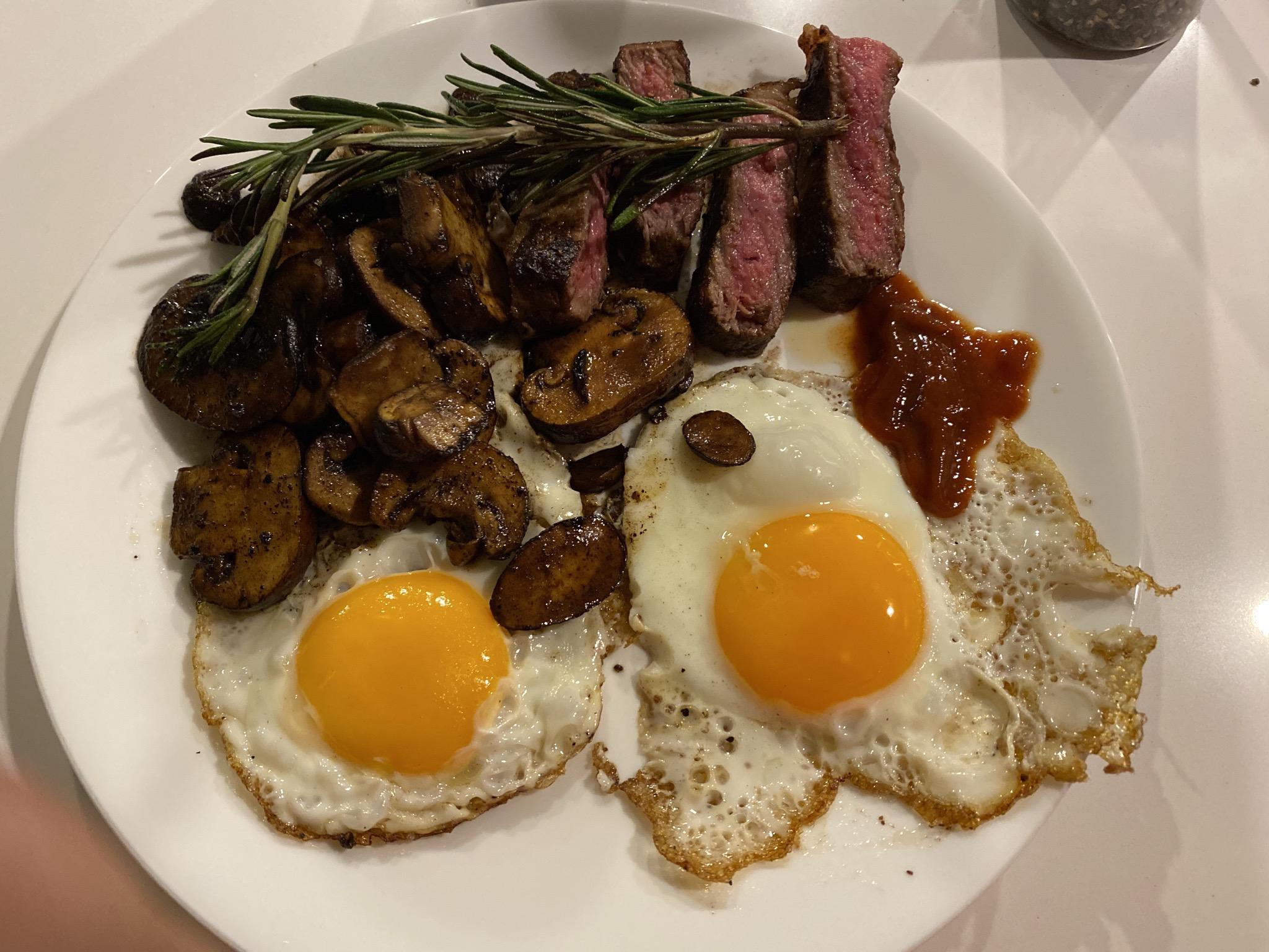 A white plate with two sunny side up eggs, sautéed mushrooms, slices of medium-rare steak topped with rosemary, and a dollop of ketchup on the side.