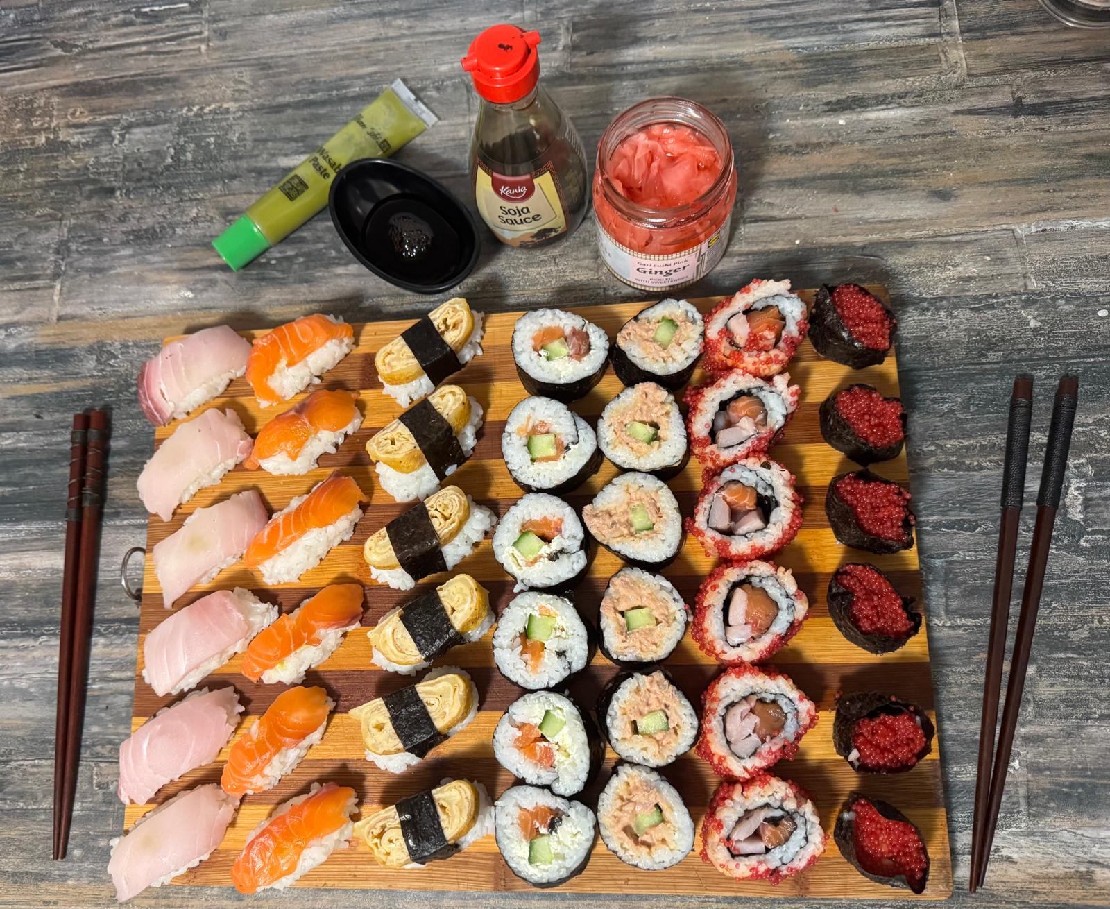 A wooden board with assorted sushi rolls, nigiri, tamago, and bright red tobiko. Soy sauce, wasabi, pickled ginger, and two pairs of chopsticks are also on the gray textured table.