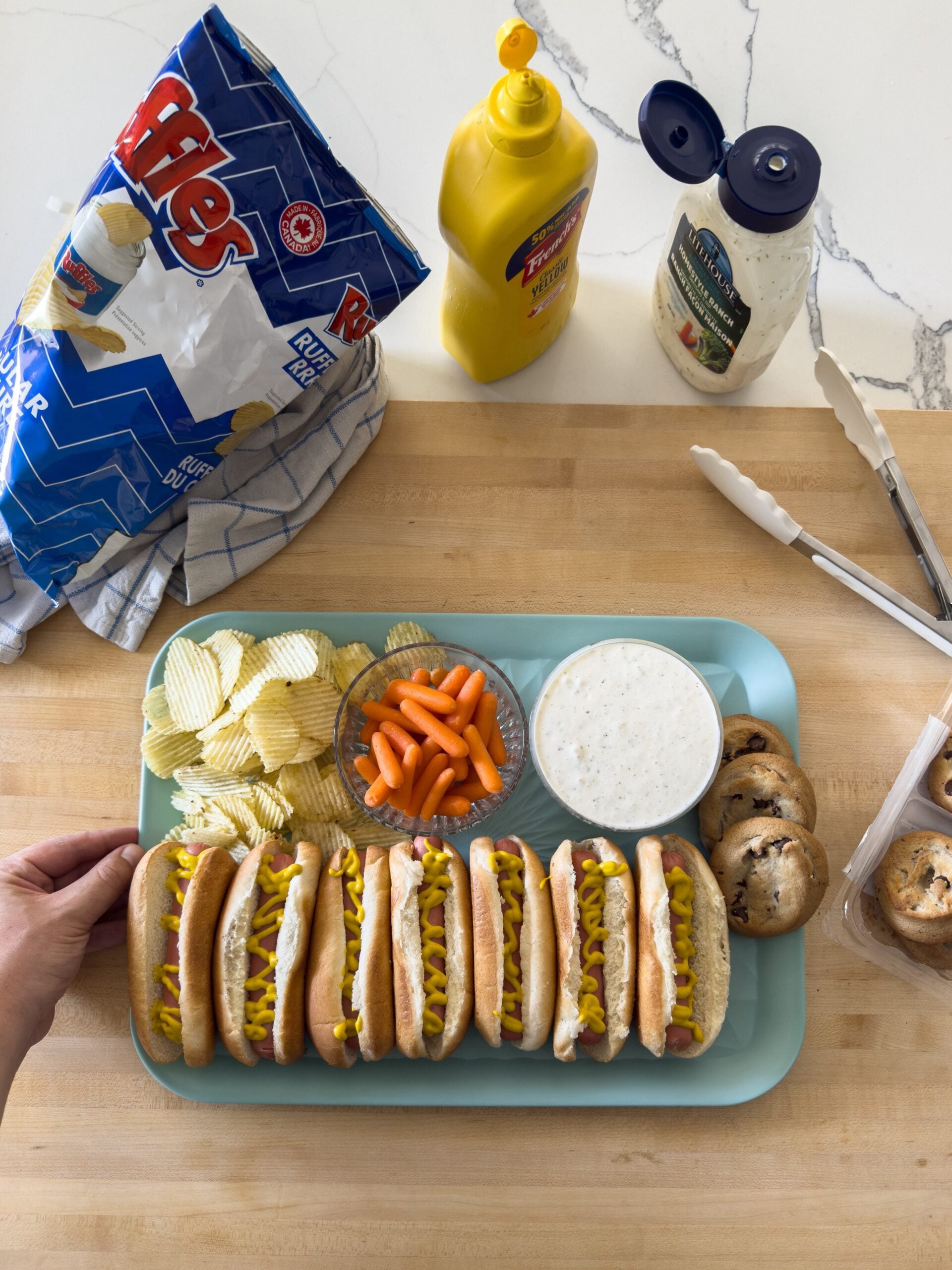 A tray with six hot dogs topped with mustard, potato chips, baby carrots, ranch dip, and chocolate chip cookies. Nearby are a bag of Ruffles chips, mustard, ranch dressing, tongs, and scissors on a wooden surface.