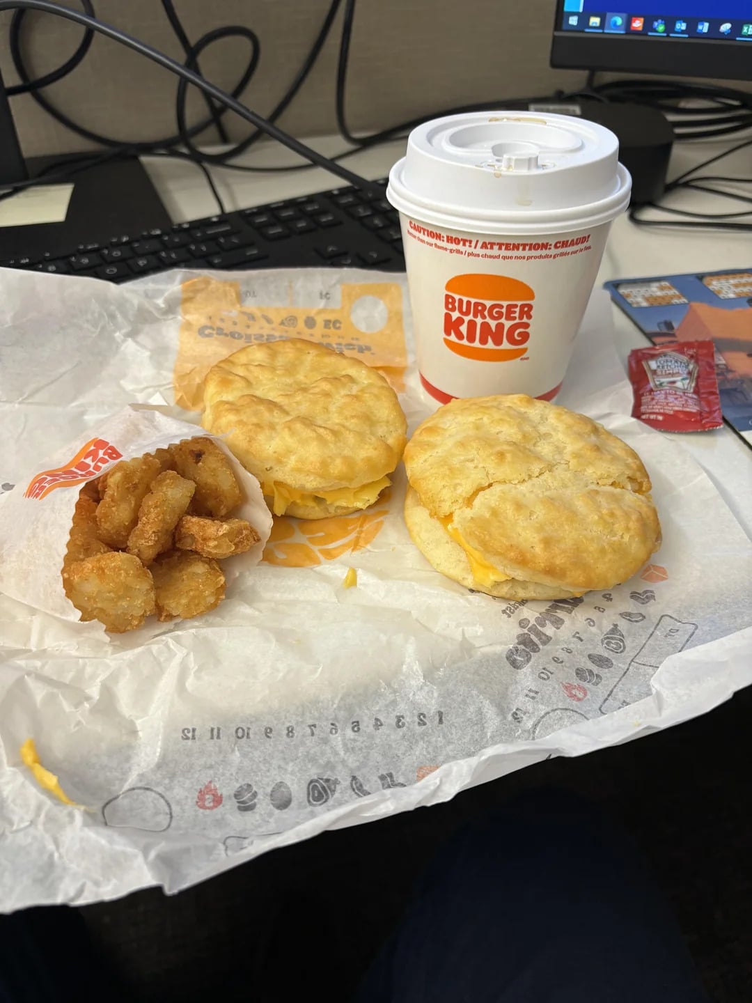Breakfast from Burger King on a desk: two egg and cheese biscuits, a small serving of hash browns, a cup of coffee, and packets of ketchup, with a keyboard and monitor in the background.