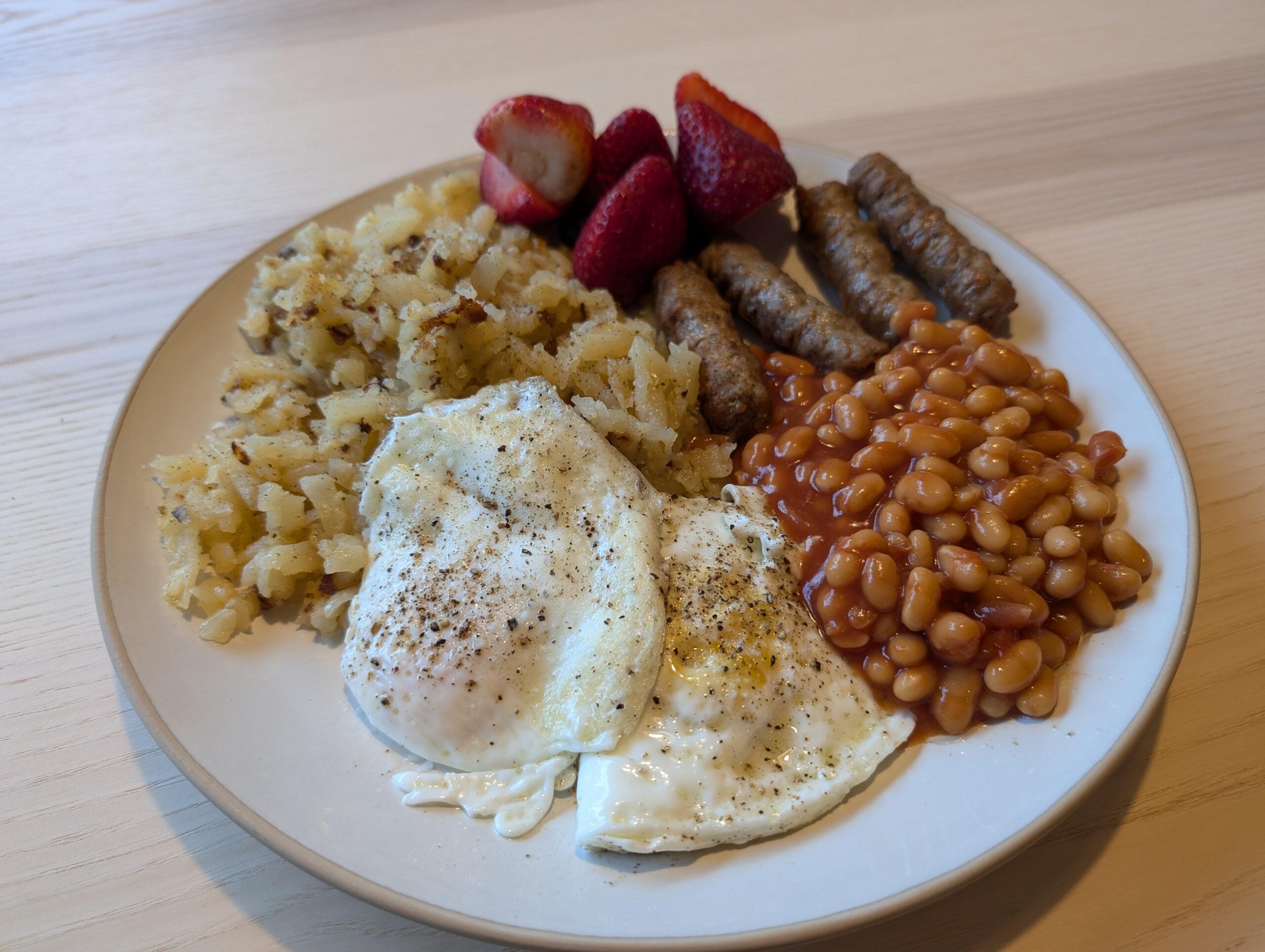 A plate with two fried eggs, baked beans, hash browns, sausage links, and sliced strawberries on a light wooden table.