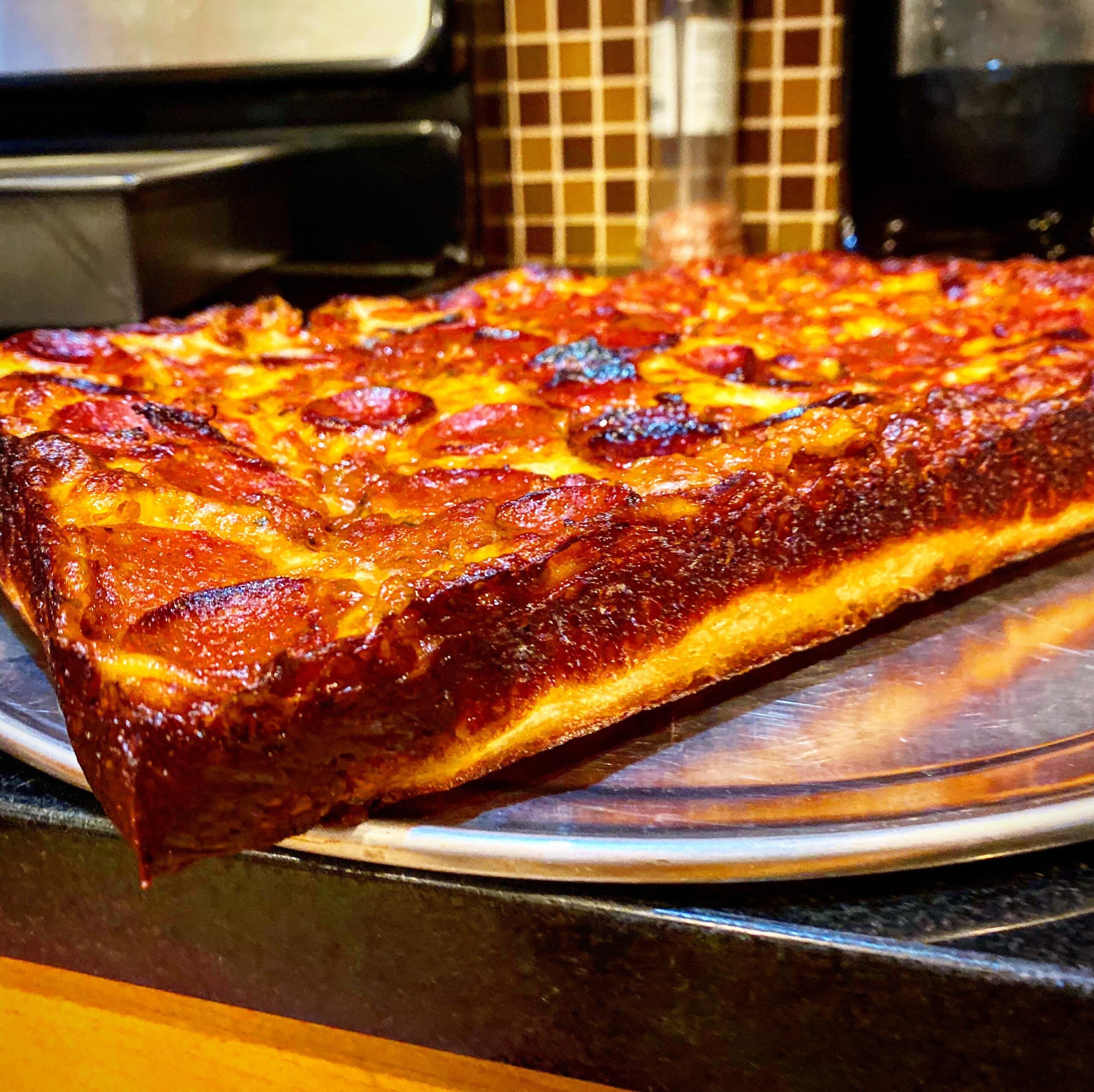 A close-up of a thick, rectangular pepperoni pizza with crispy, caramelized edges sits on a metal tray in a kitchen.