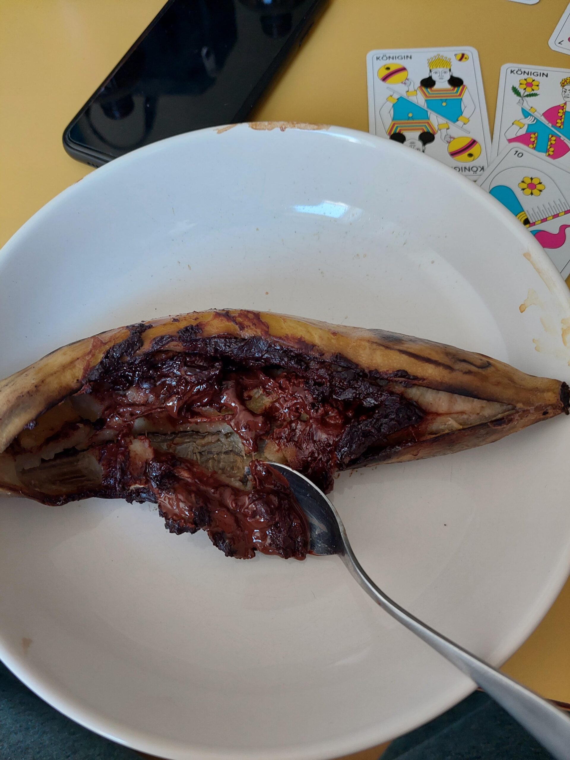 A baked banana split open in a white bowl, filled with melted chocolate. A spoon rests in the bowl. Playing cards and a phone are visible on the yellow table in the background.